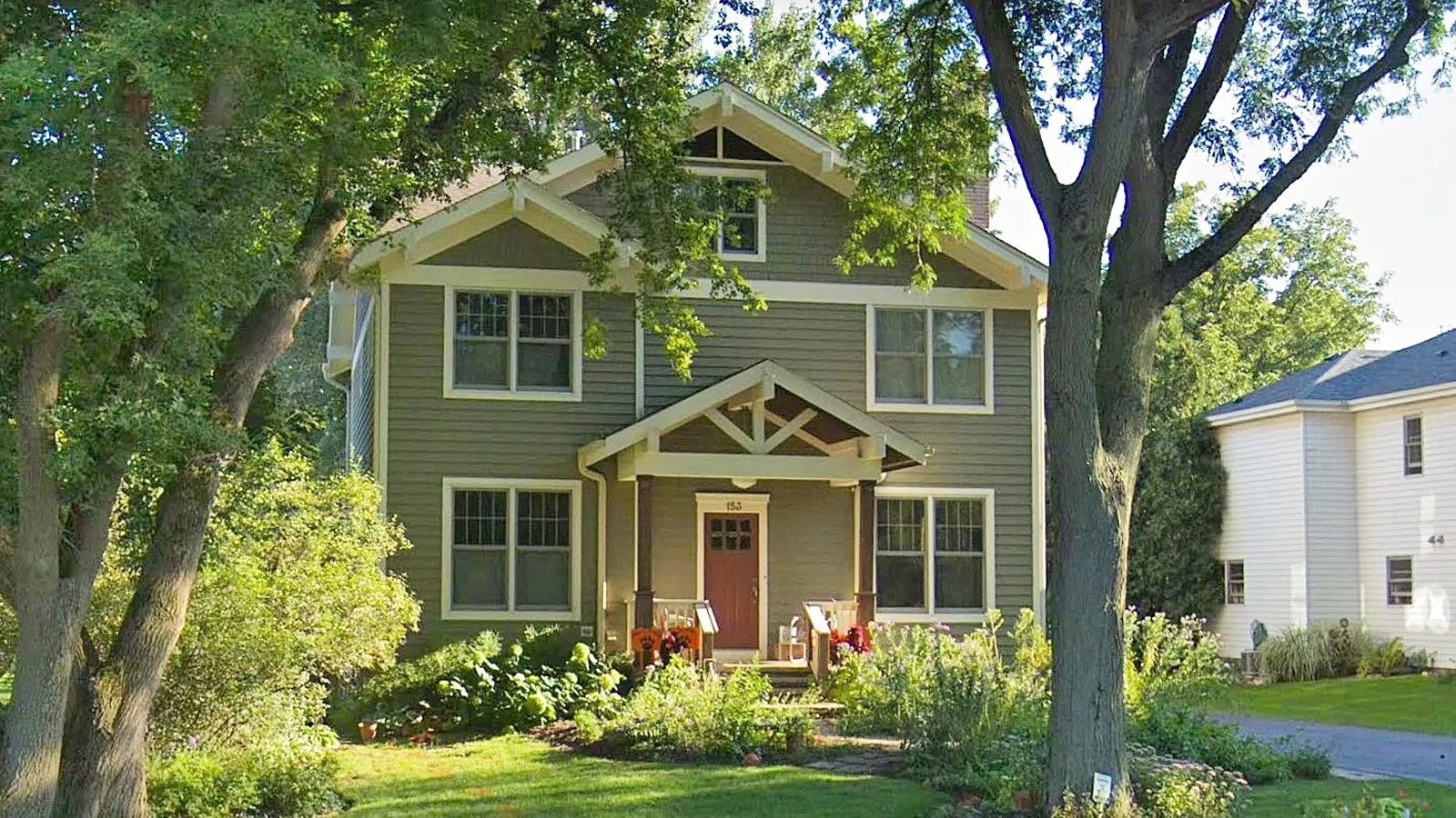 Two-story green house with white trim and a front porch, surrounded by trees and a sunny yard.