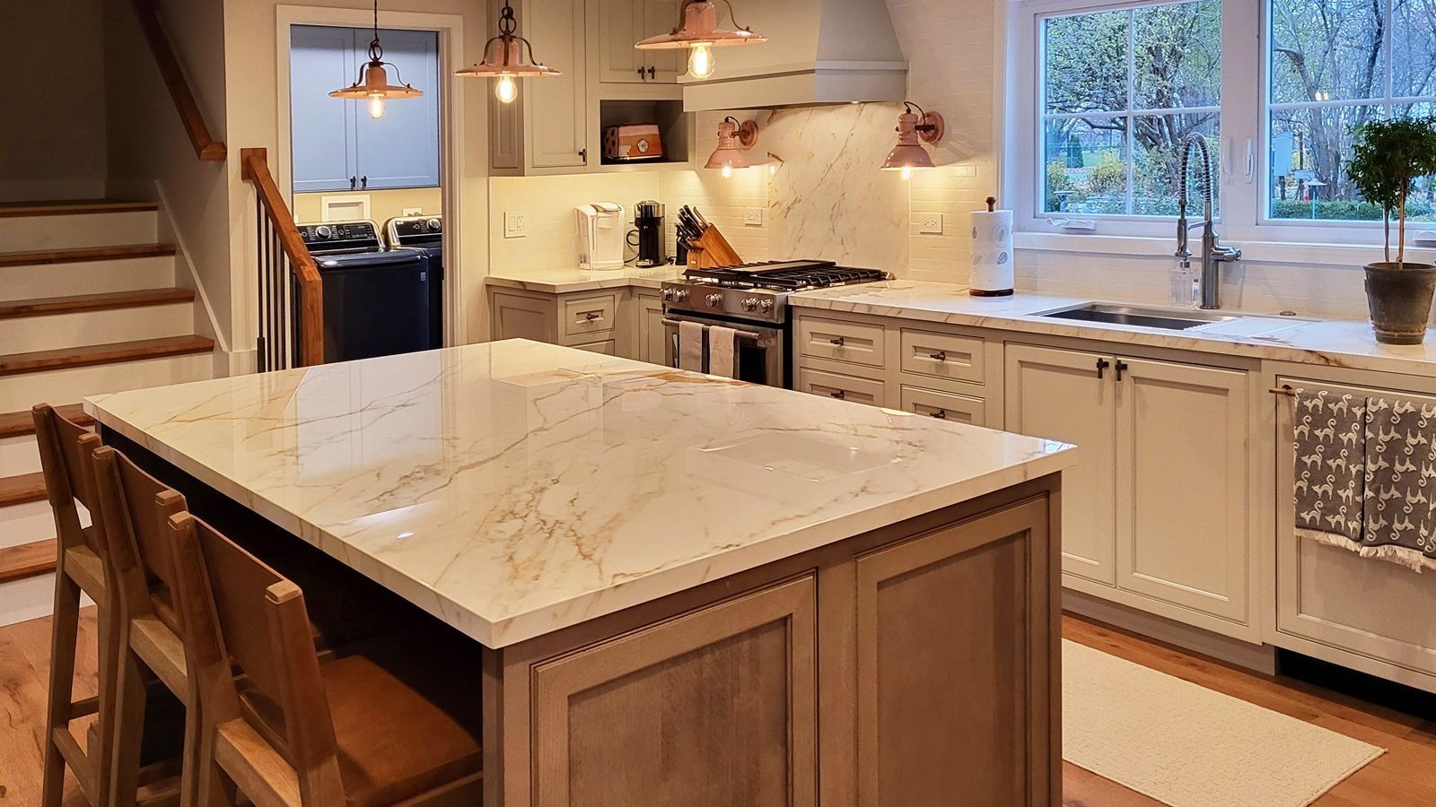 Bright modern kitchen with a marble island, wooden stools, white cabinets, and a sunlit window