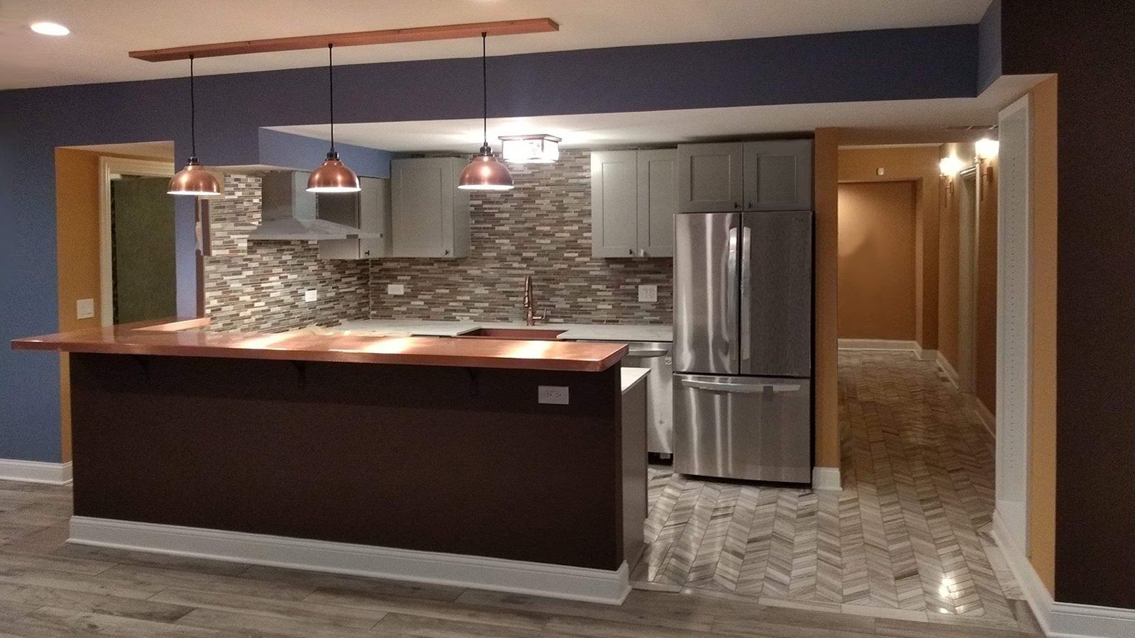 Modern kitchen with dark island, stainless steel refrigerator, pendant lights, and stone tile floor