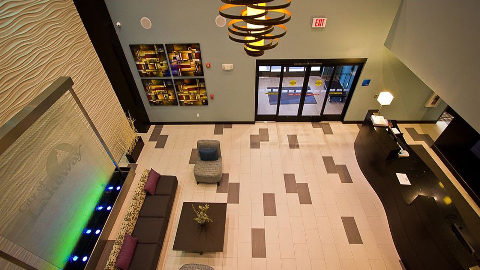 Lobby with tan tile floor, dark chairs, wall art, and glass entrance doors under modern ceiling lights