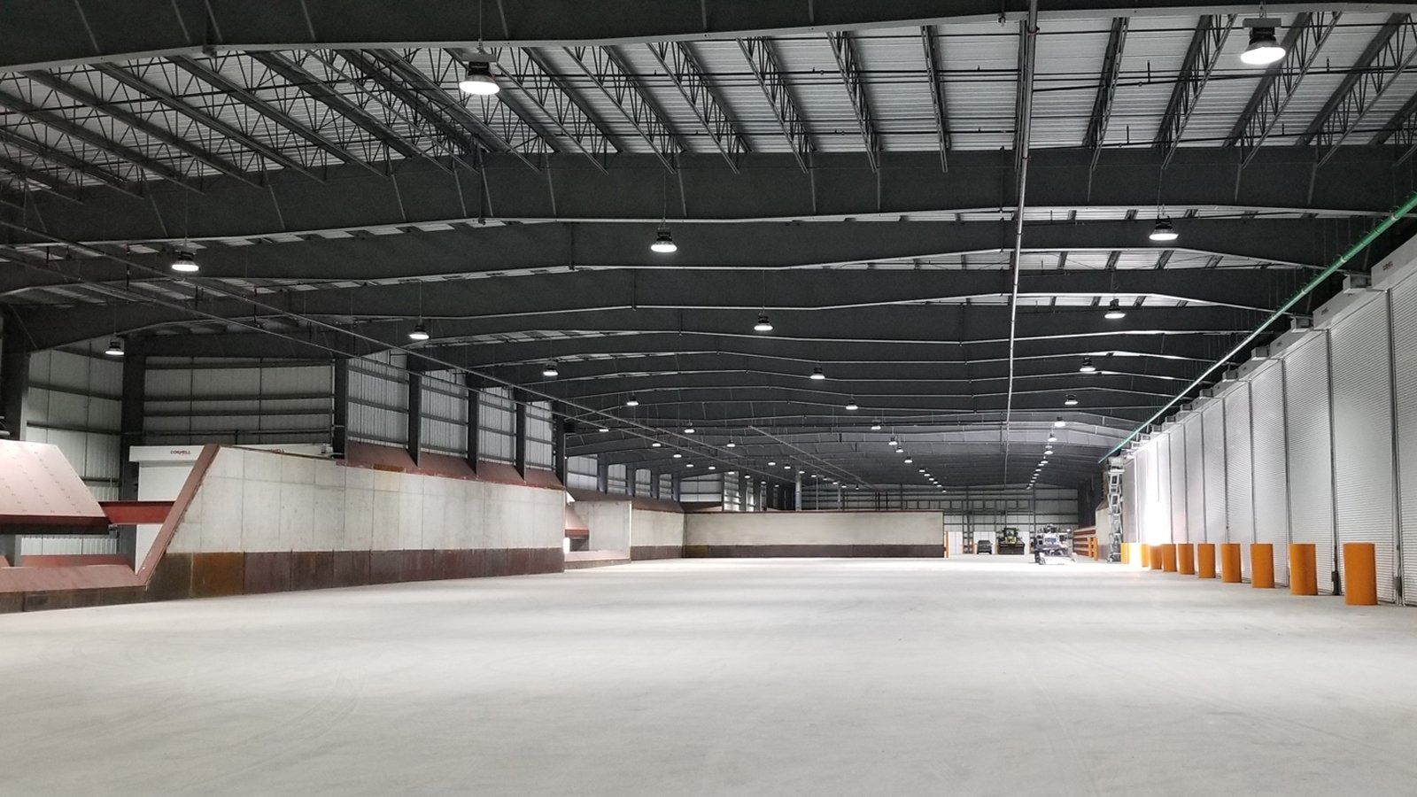 Empty indoor warehouse with a high metal roof and concrete floor, lit by skylights and side windows