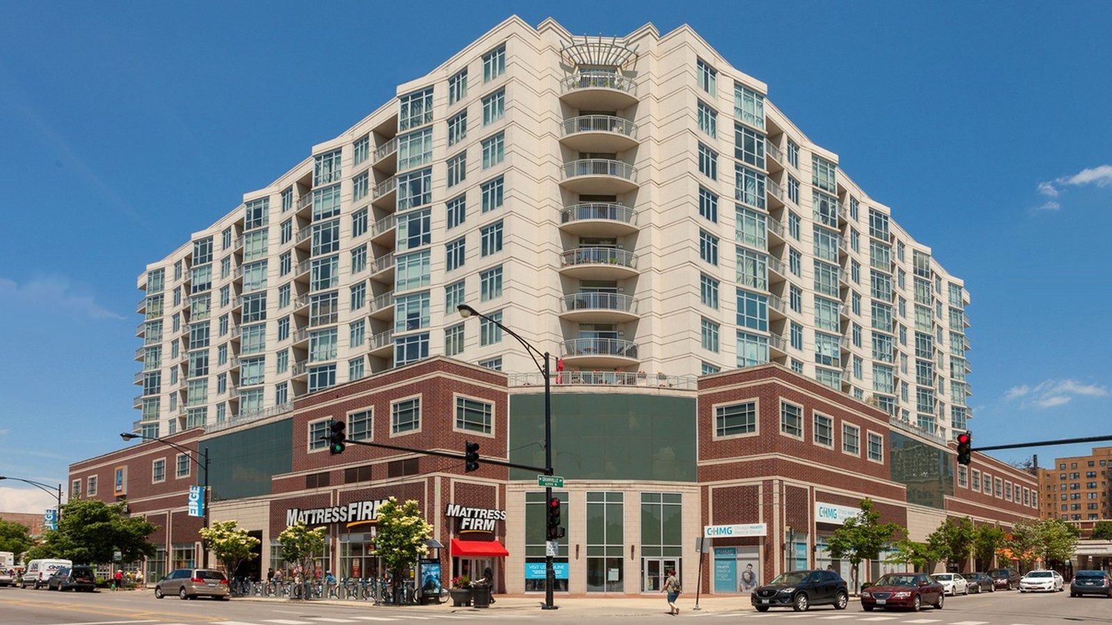 Street corner with a red-brick and beige high-rise apartment building under a clear blue sky.