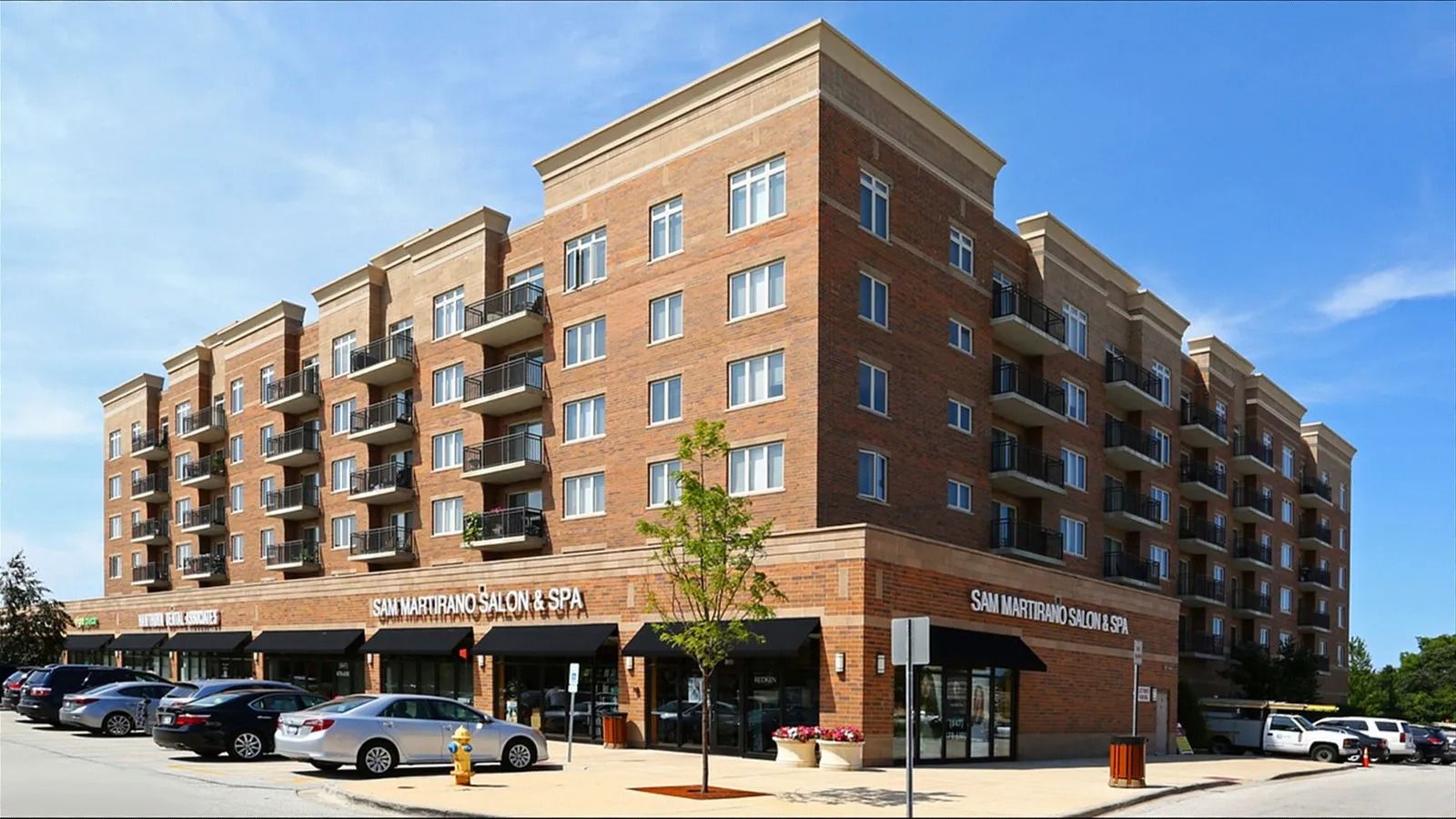 Brick apartment building with ground-floor shops and parked cars on a sunny street corner