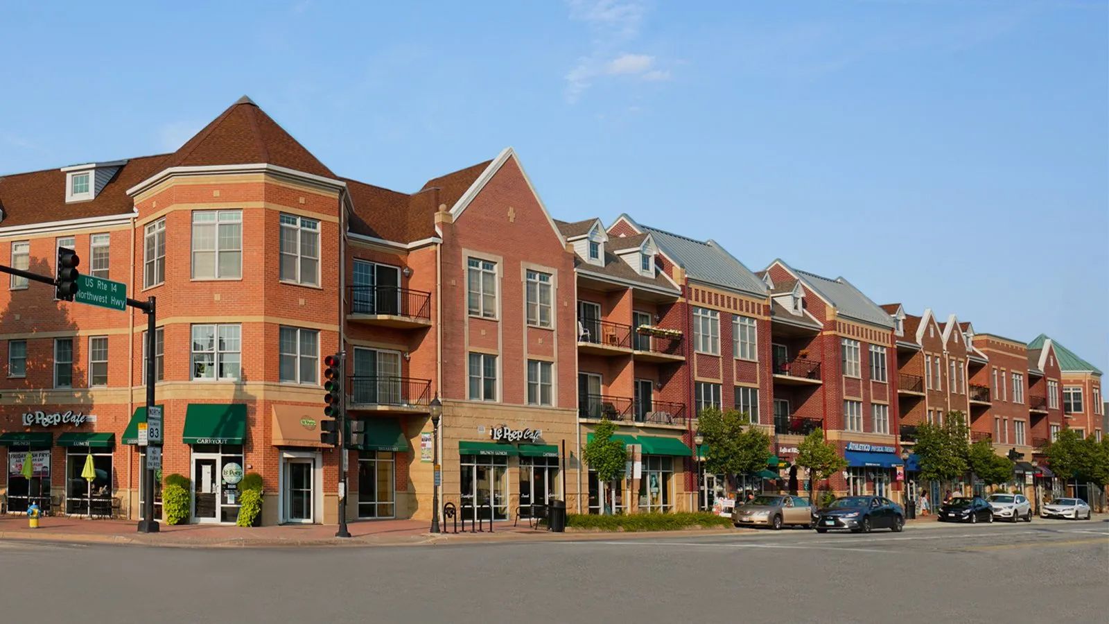 Row of red-brick storefront apartments on a sunny street corner with parked cars and trees