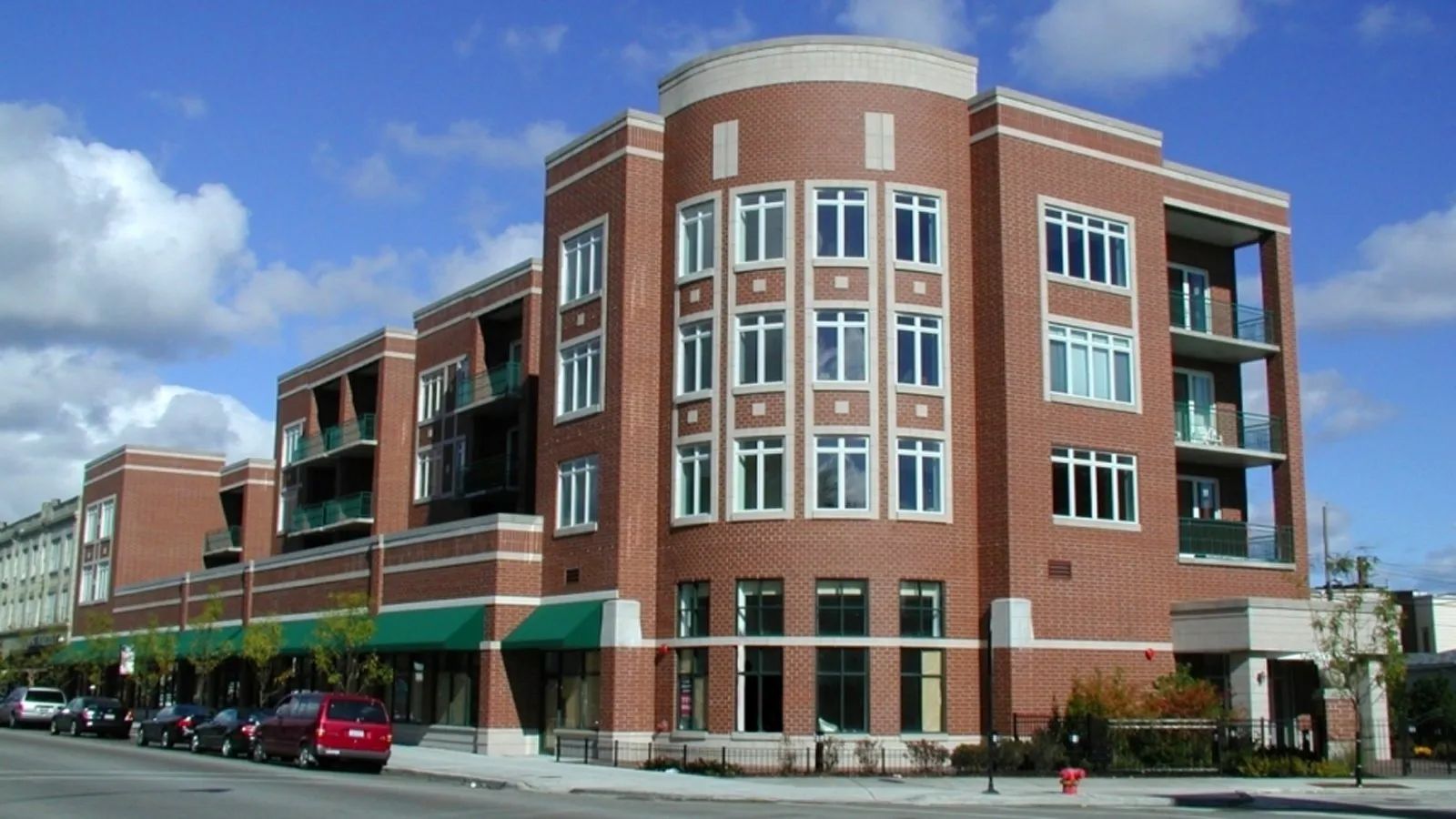 Modern brick apartment building on a sunny street corner with green awnings and parked cars