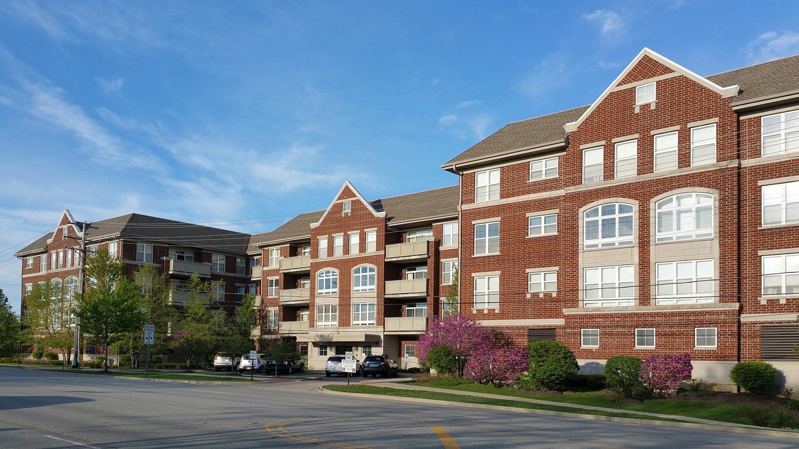 Brick apartment buildings with white trim and blooming landscaping beside an empty parking lot