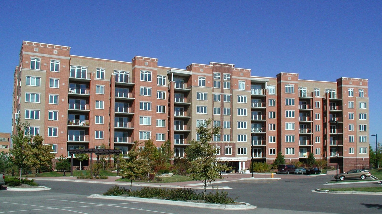 Brick apartment building with many windows and balconies beside a parking lot under a clear sky