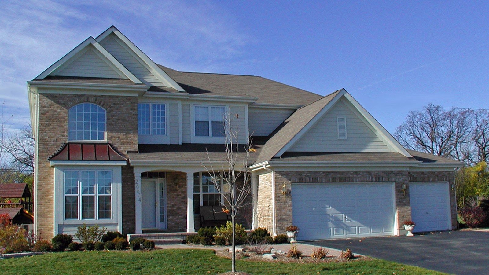 Two-story suburban house with beige siding, stone accents, and a two-car garage under a blue sky