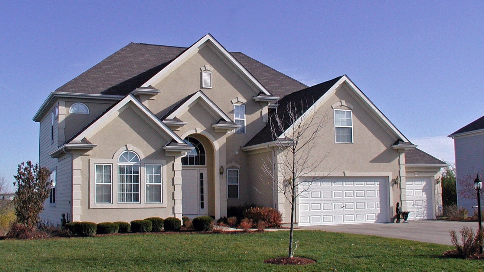 Suburban two-story beige house with a gray roof, white garage, and front lawn under a clear sky