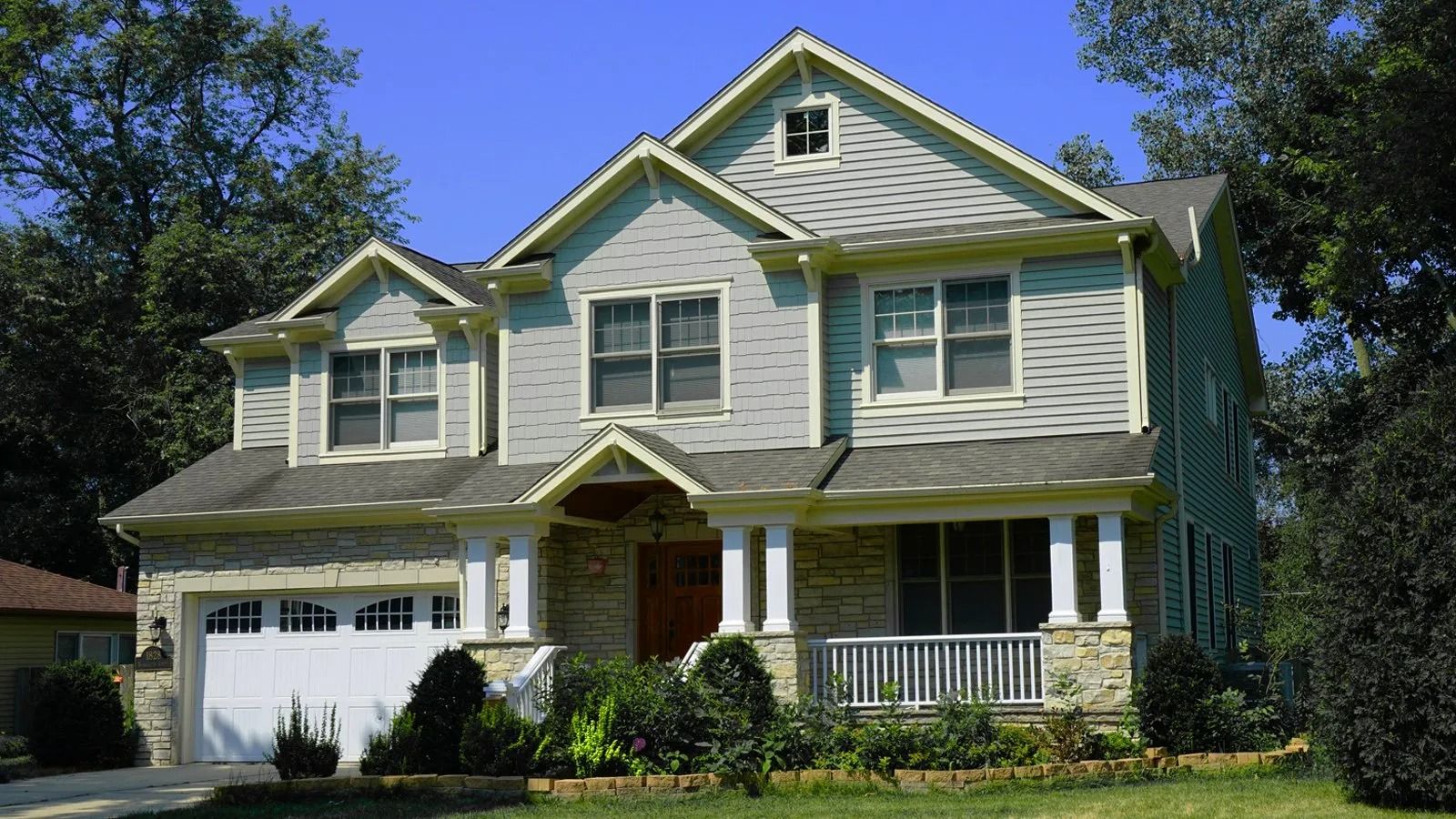 Two-story suburban house with a garage, white siding, green shutters, and a front porch under blue sky