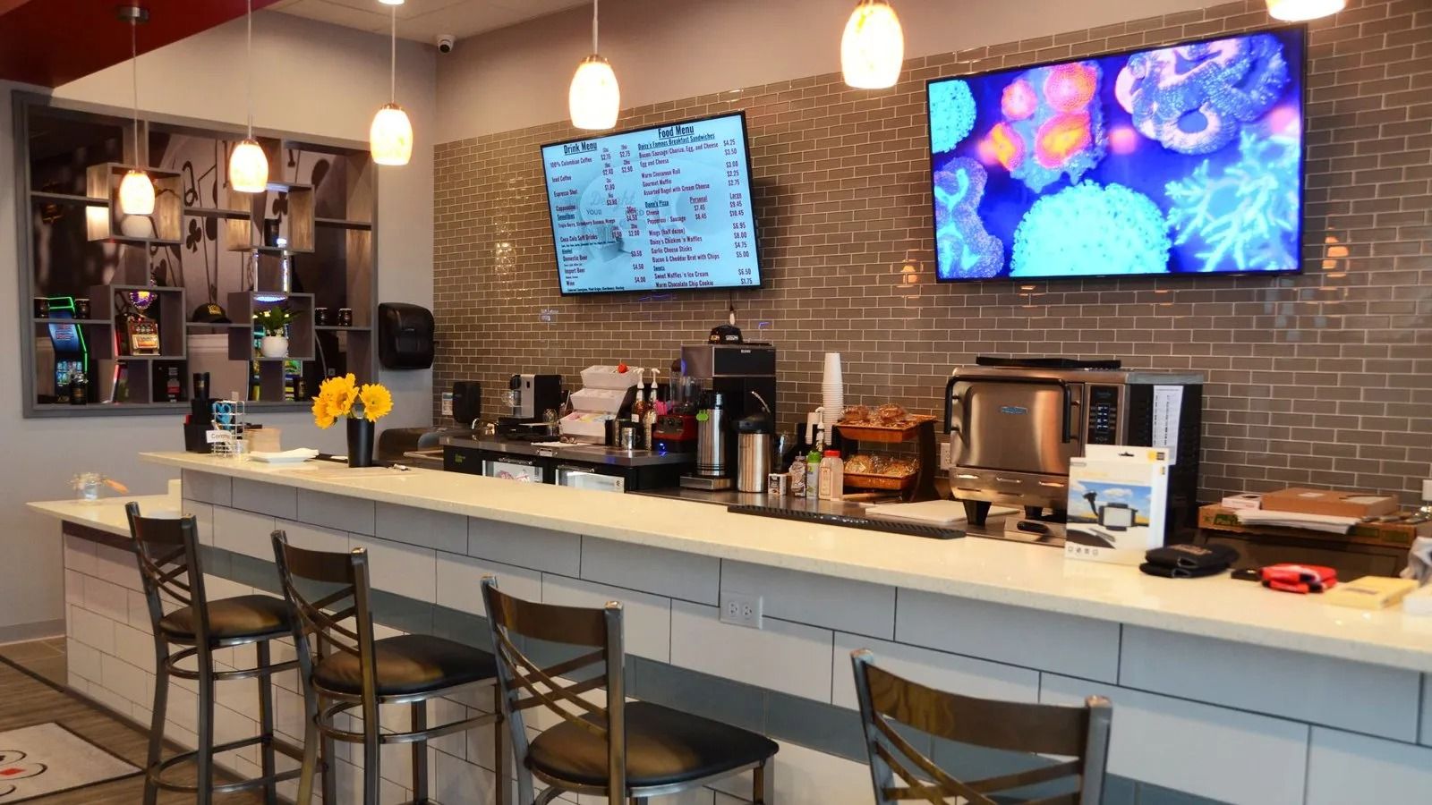 Modern café counter with stools, pendant lights, brick wall, and two wall-mounted screens.