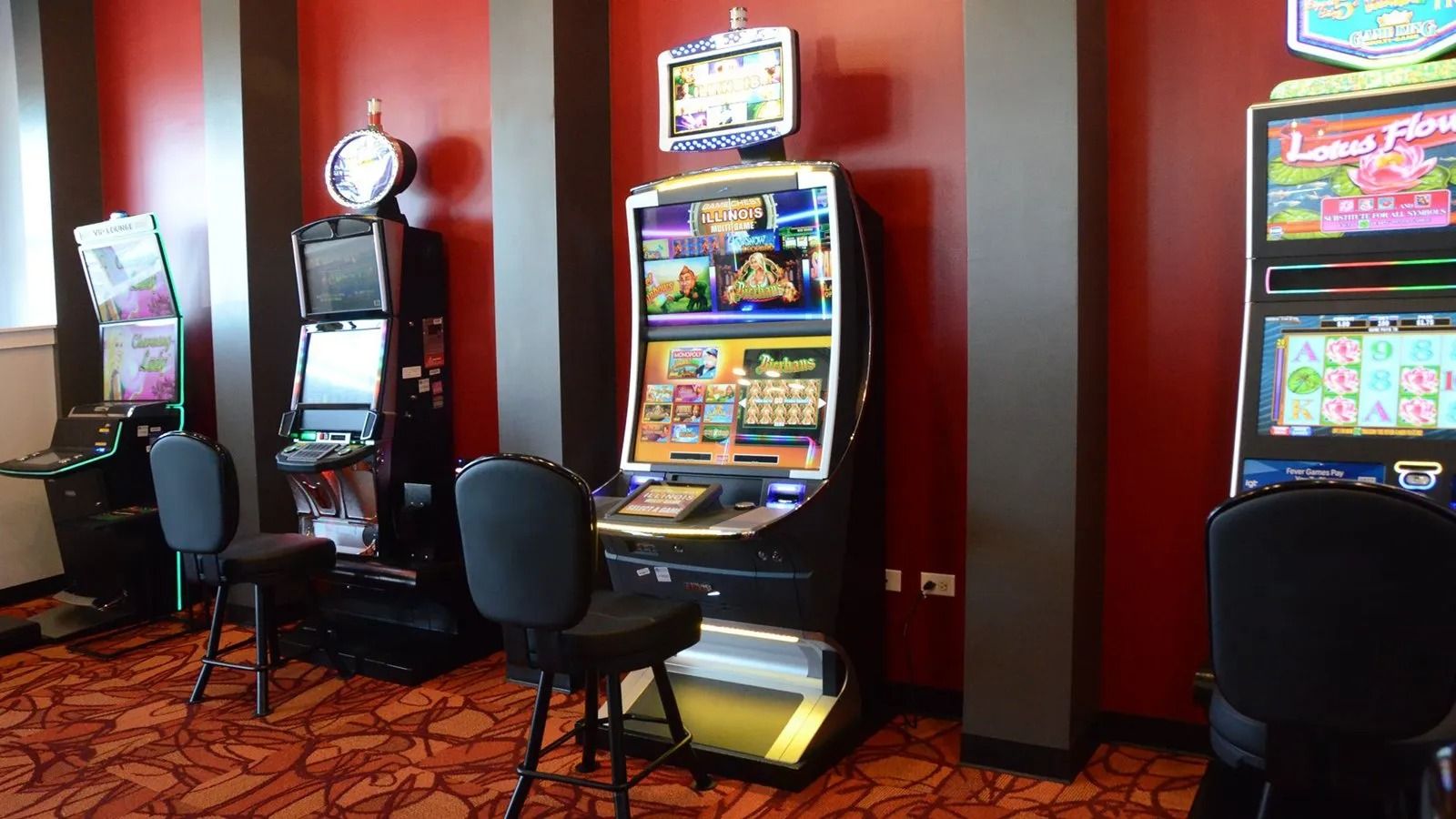 Row of colorful slot machines in a casino with black chairs and patterned red carpet.