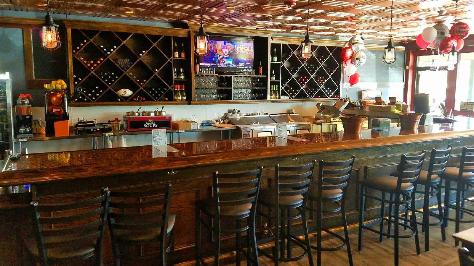 Empty bar interior with wooden stools, long counter, shelves of bottles, and hanging red lanterns