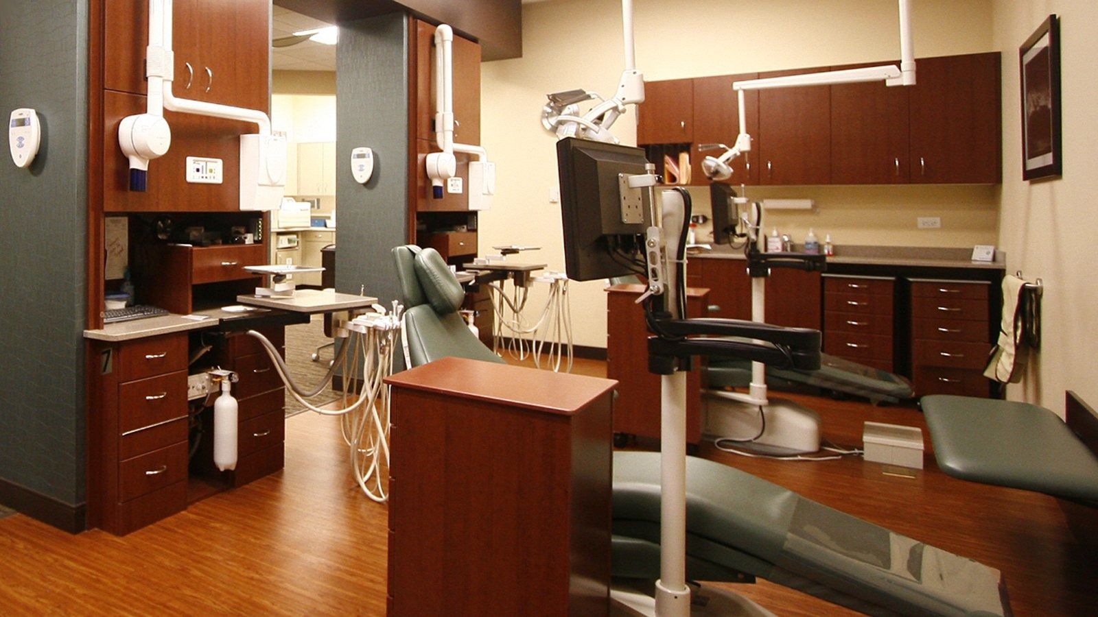 Empty dental exam room with chairs, cabinets, and overhead lights in a wood-floored clinic.
