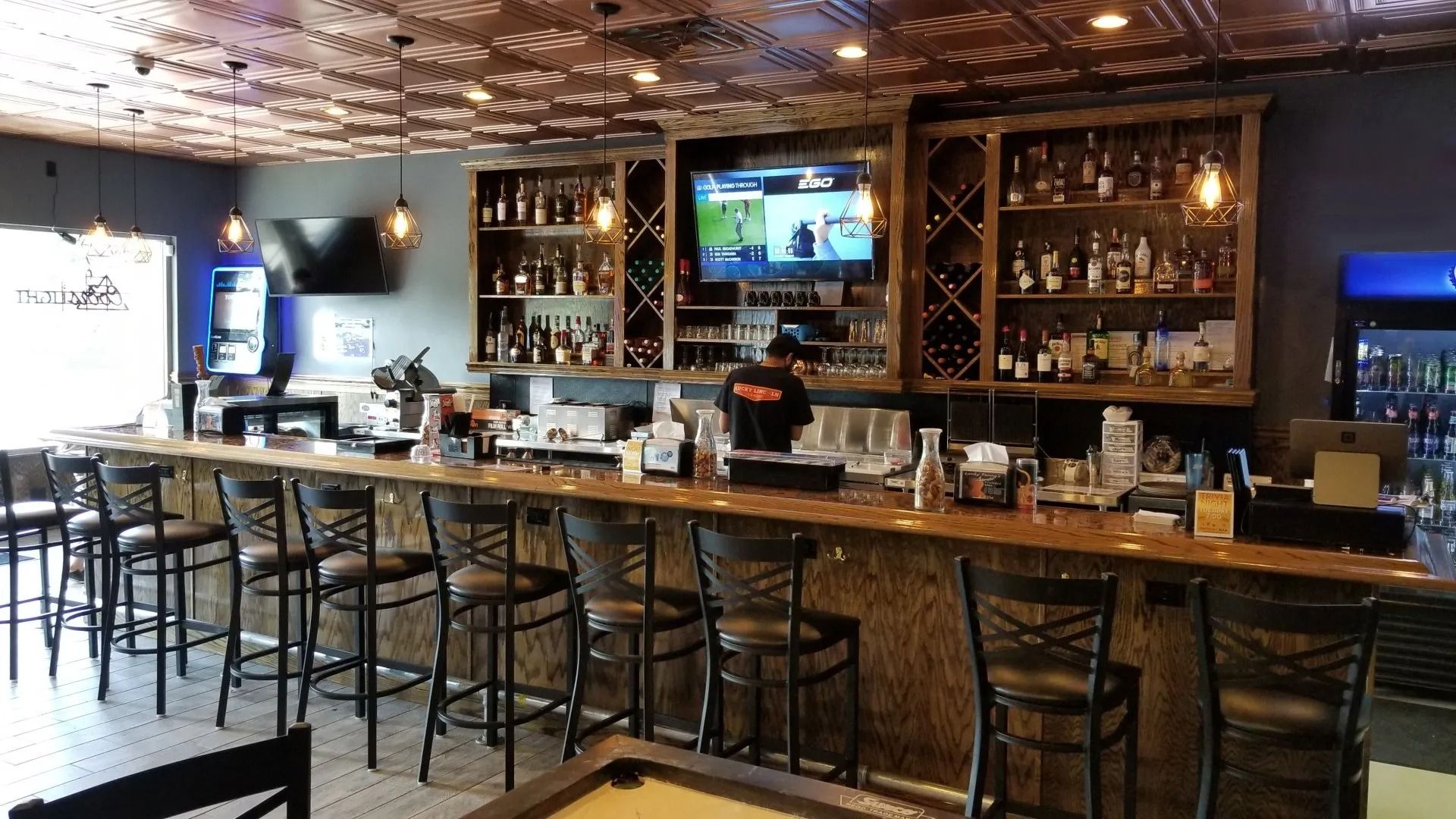 Empty bar interior with stools along a wooden counter, wine shelves, and TV screens on the back wall