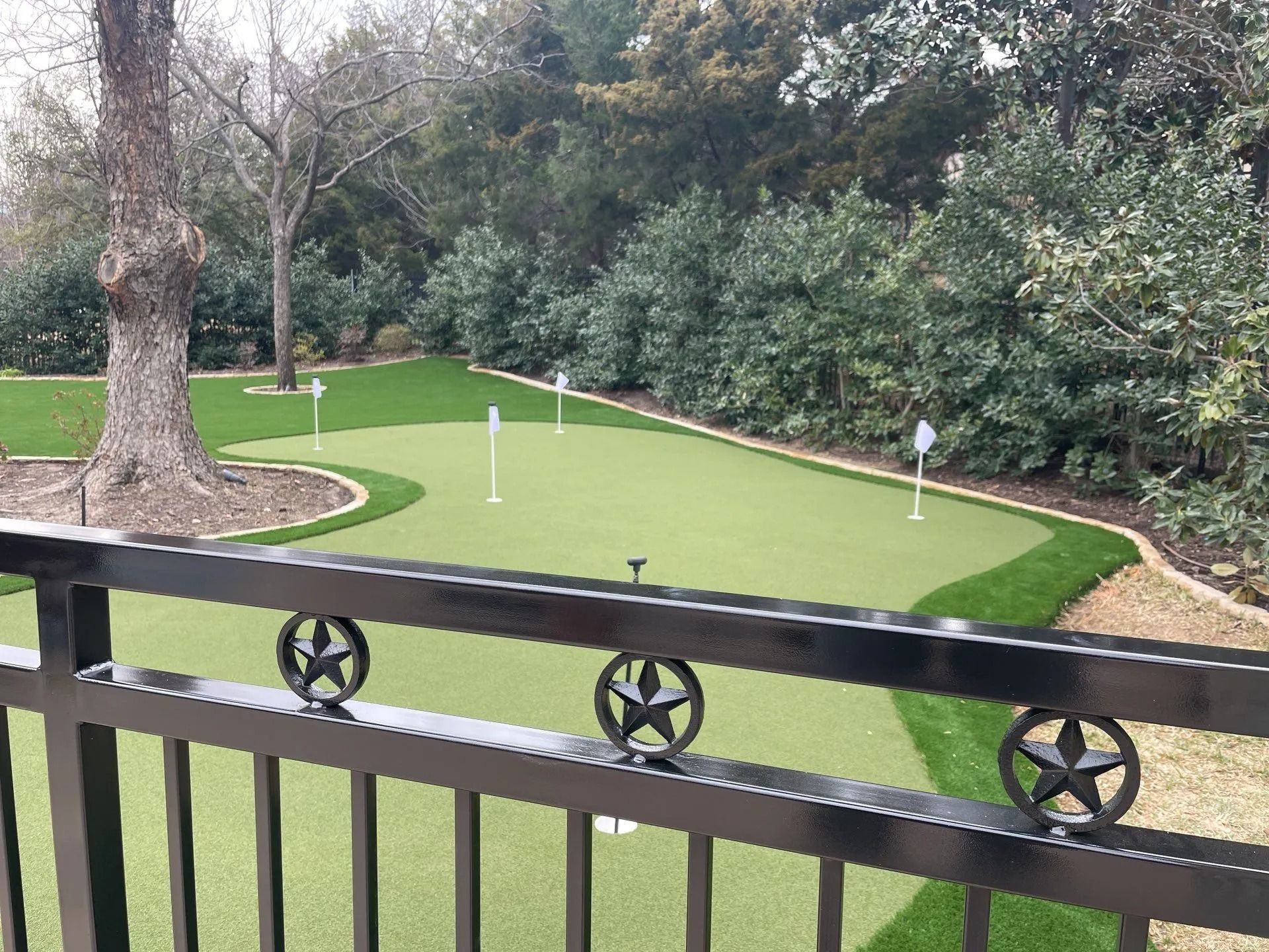 Small putting green in a park, viewed from a balcony with black railing, surrounded by trees and shrubs.