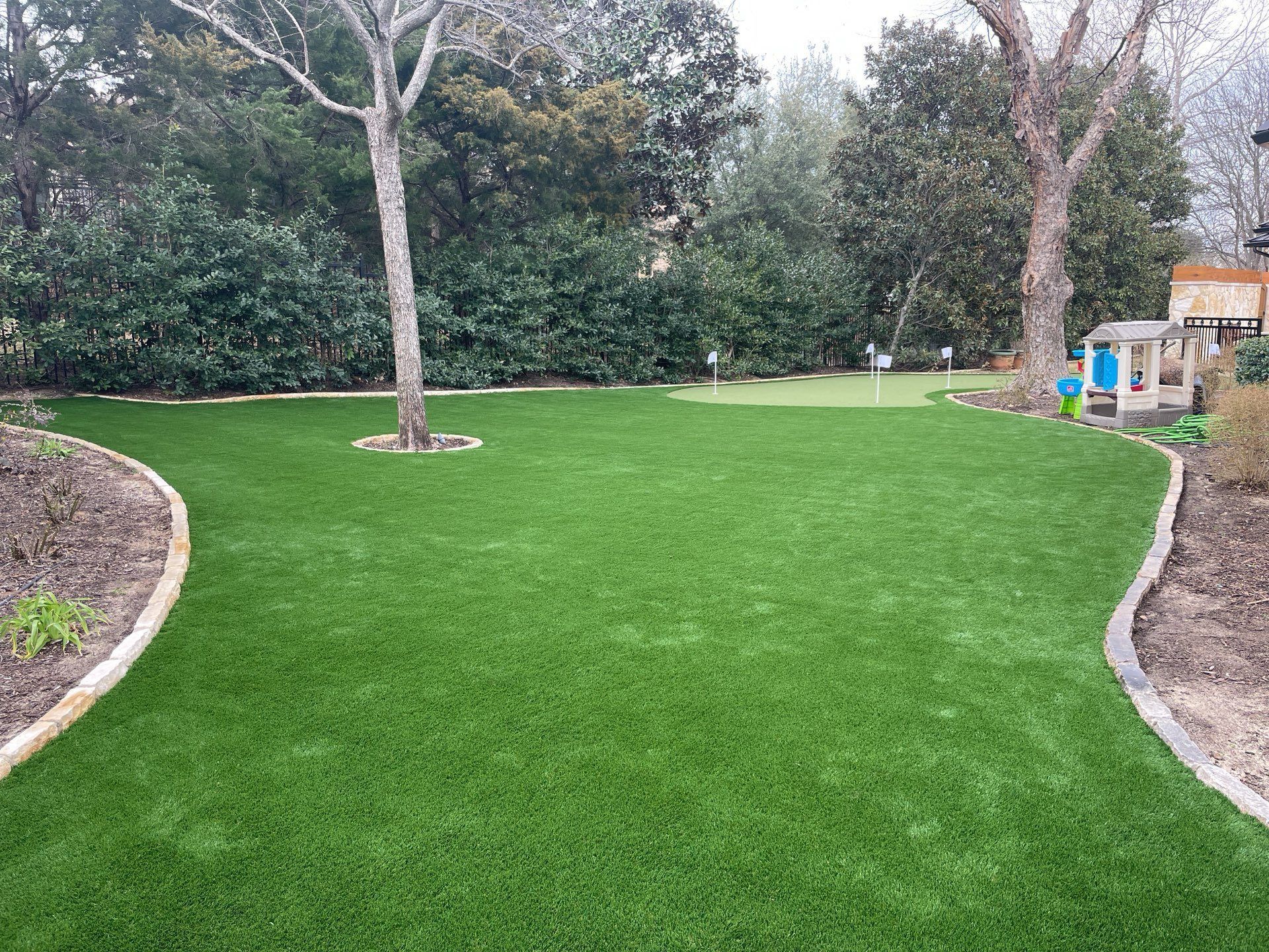 Green lawn in a landscaped yard with trees, shrubs, and a small playhouse on the right.