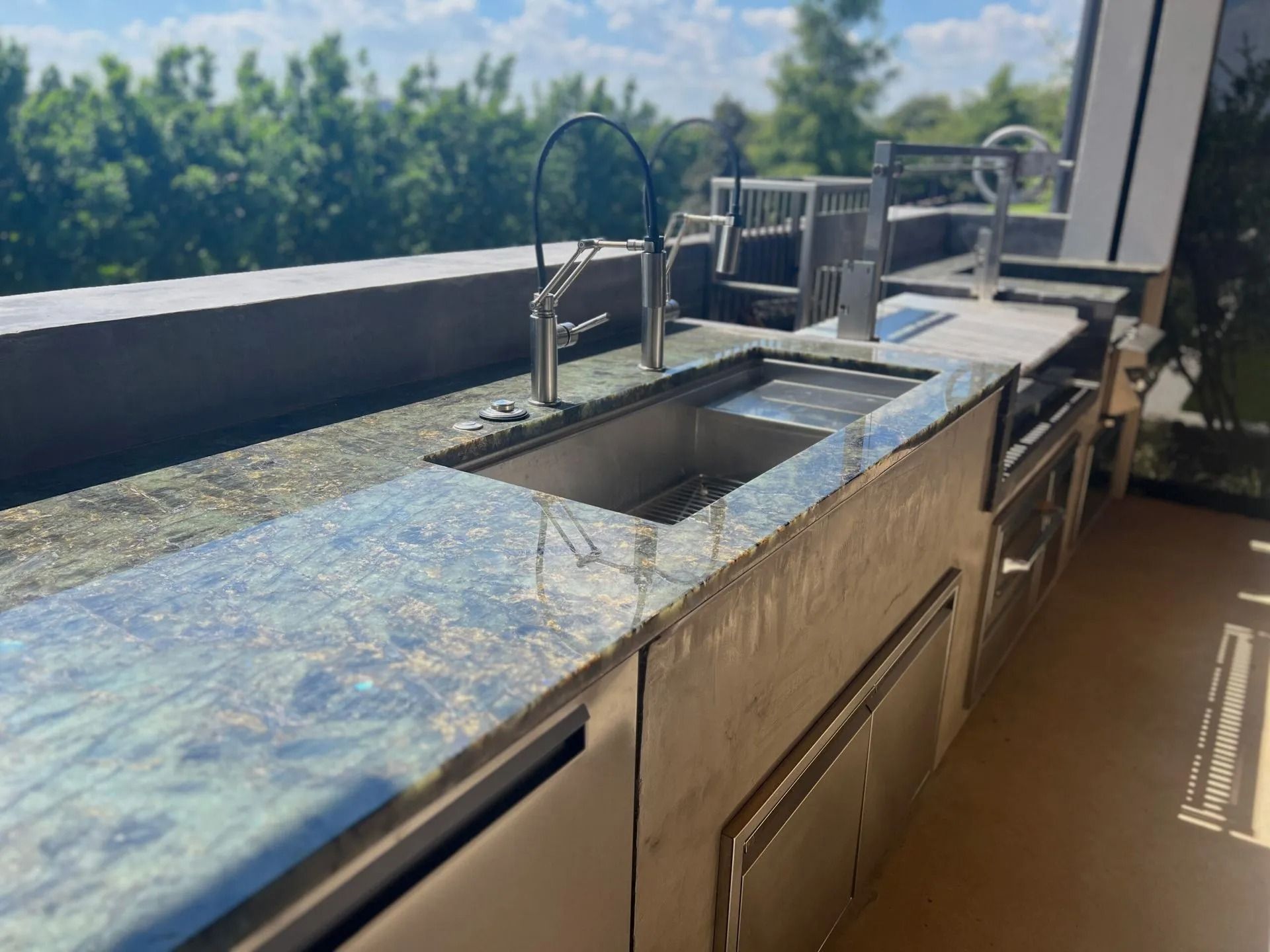 Outdoor granite countertop with stainless sink and faucet by a pool, overlooking trees and sky