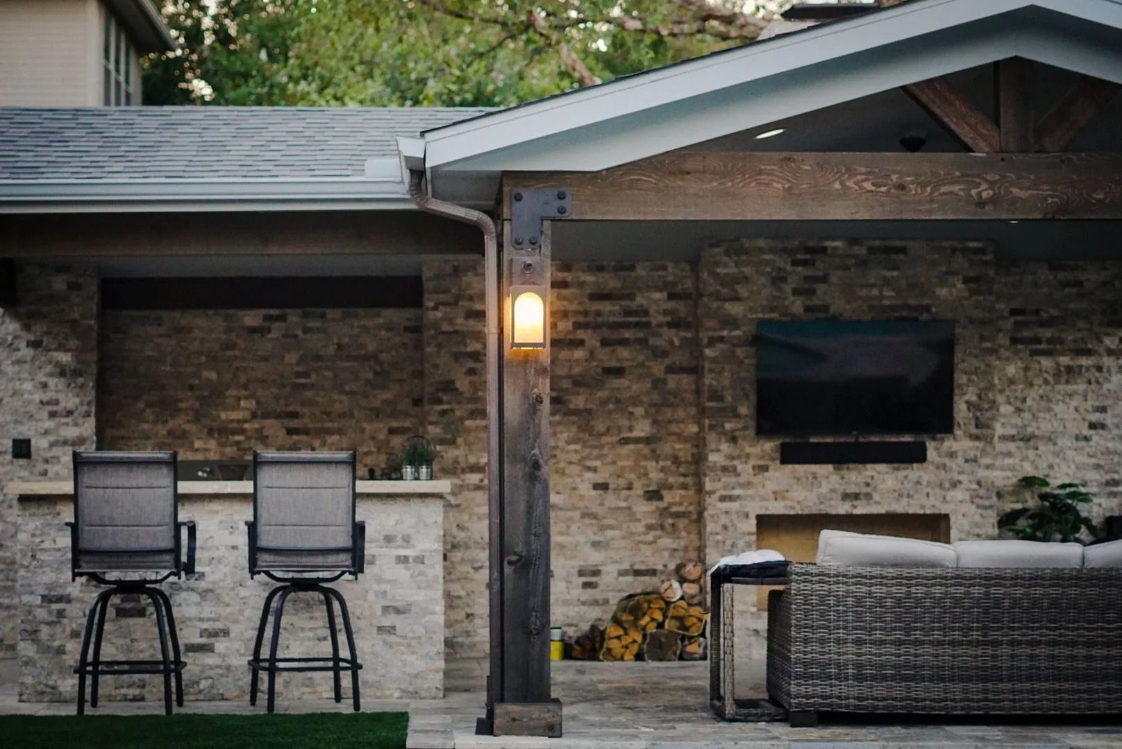 Covered backyard patio with bar stools, wall-mounted TV, and wicker sofa under a lantern.