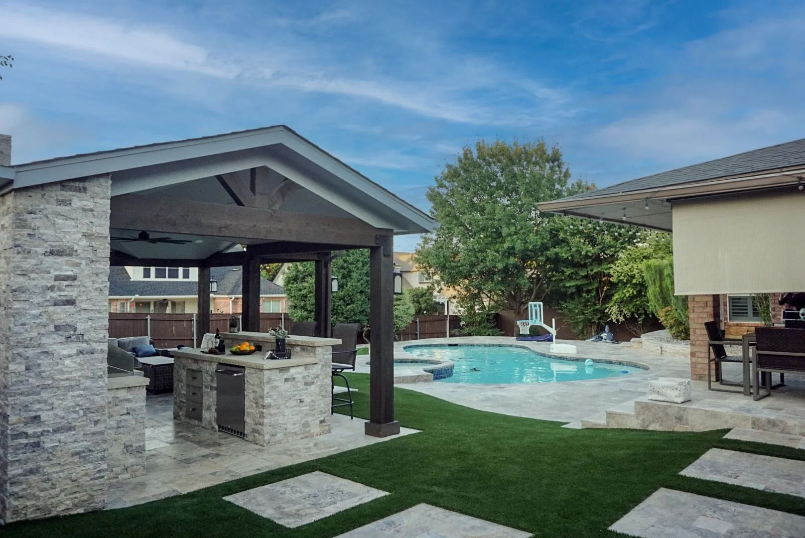 Backyard patio with pool, outdoor kitchen, stone pavilion, and lawn under a blue sky
