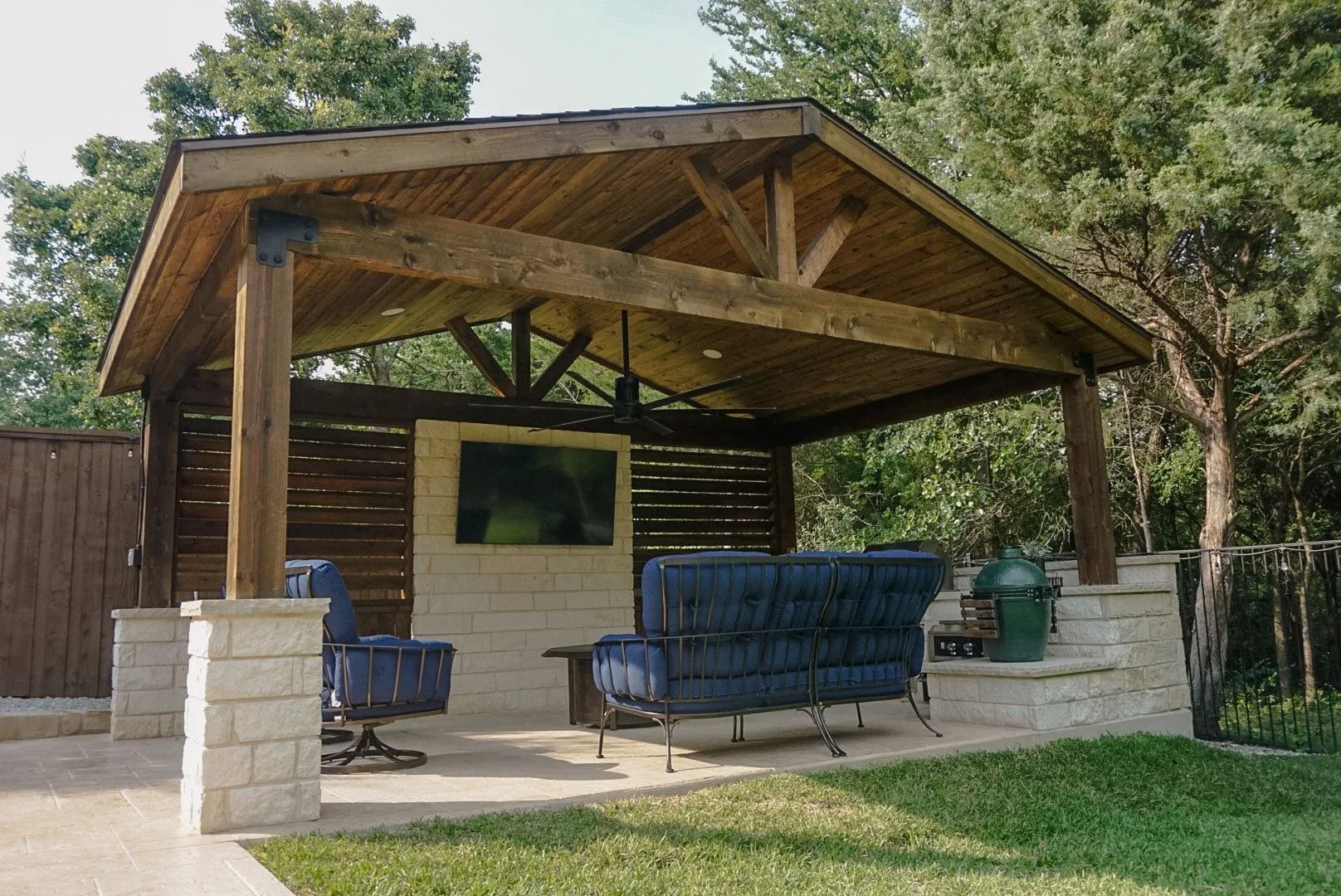 Wooden backyard pavilion with cushioned seating, stone pillars, and a small wall-mounted TV.
