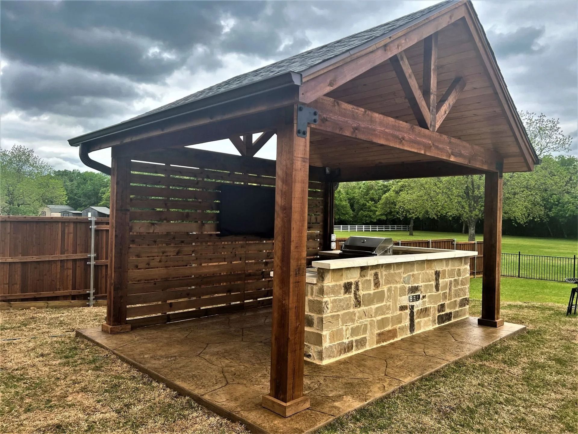 Wooden outdoor pavilion with a stone bar under a sloped roof in a grassy backyard.