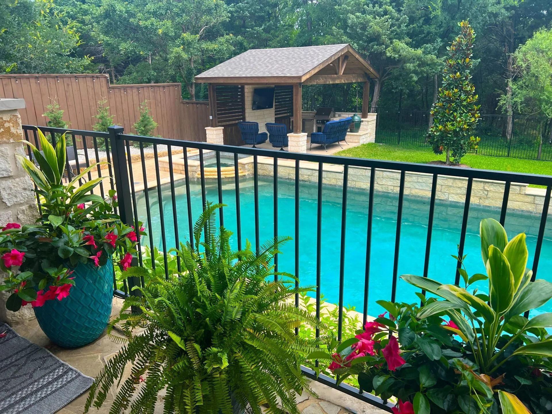 Backyard pool with black fence, potted plants, and a wooden gazebo beside a lush green lawn
