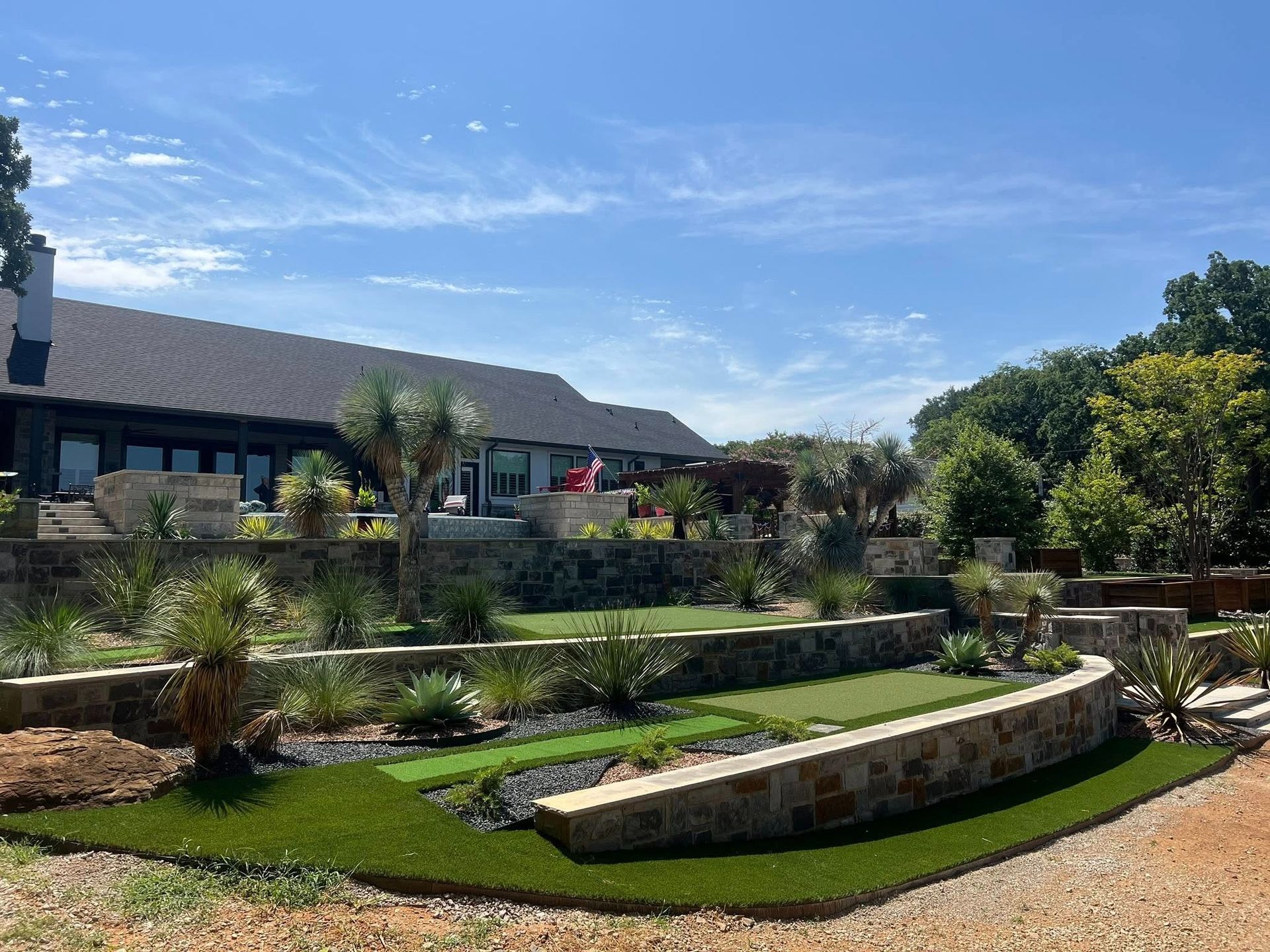 Landscaped garden with stone retaining walls, green lawn, and a long house under a blue sky