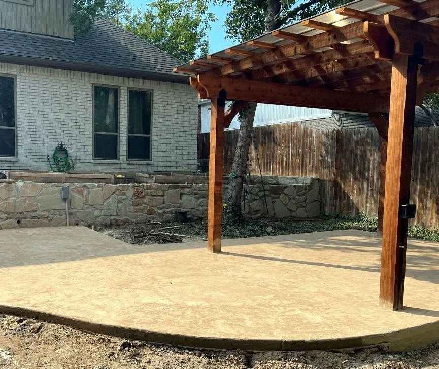 Covered patio with wooden pergola beside a stone house and concrete patio area
