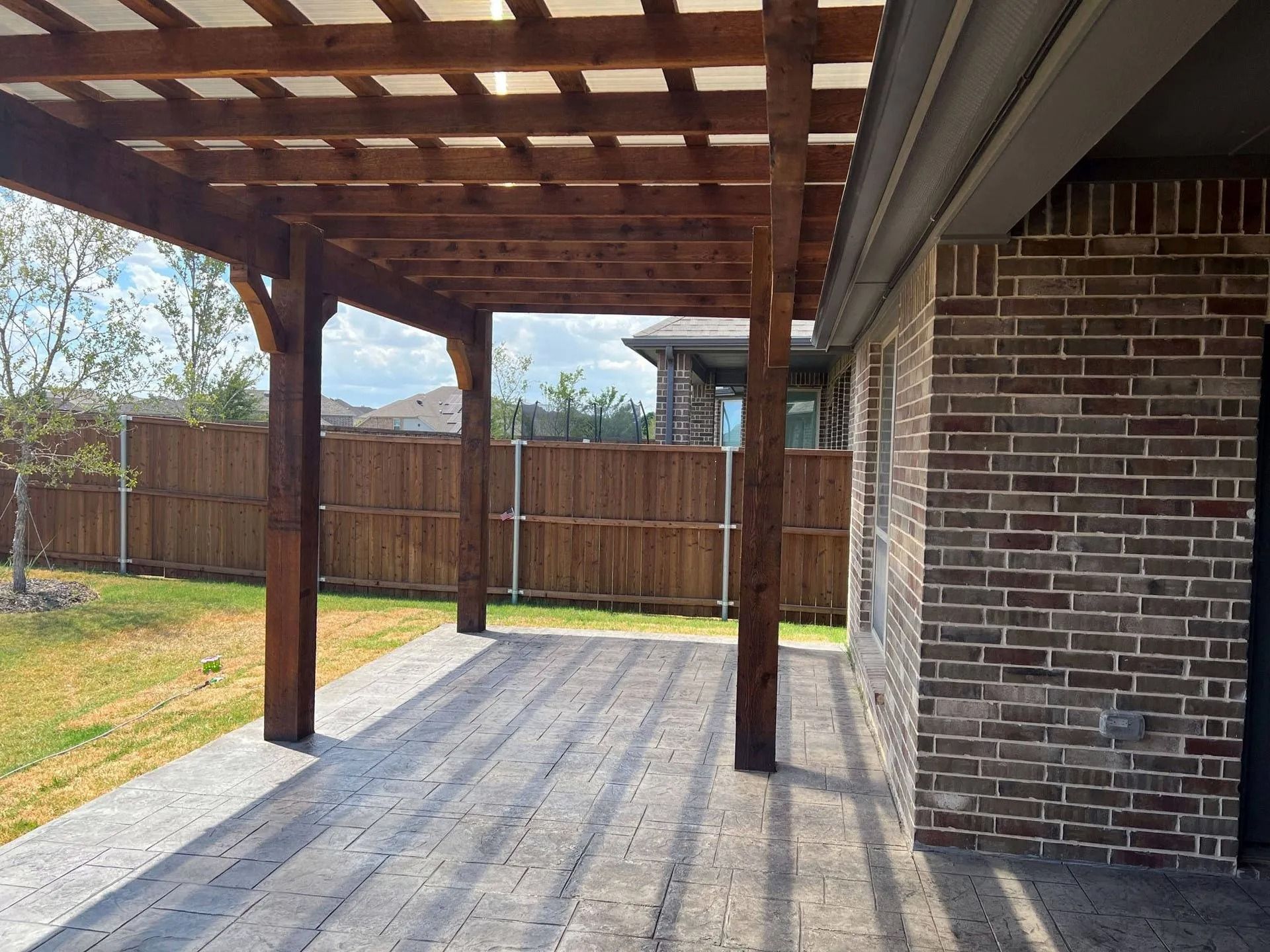 Covered backyard patio with wooden pergola, brick wall, and fenced yard in sunlight
