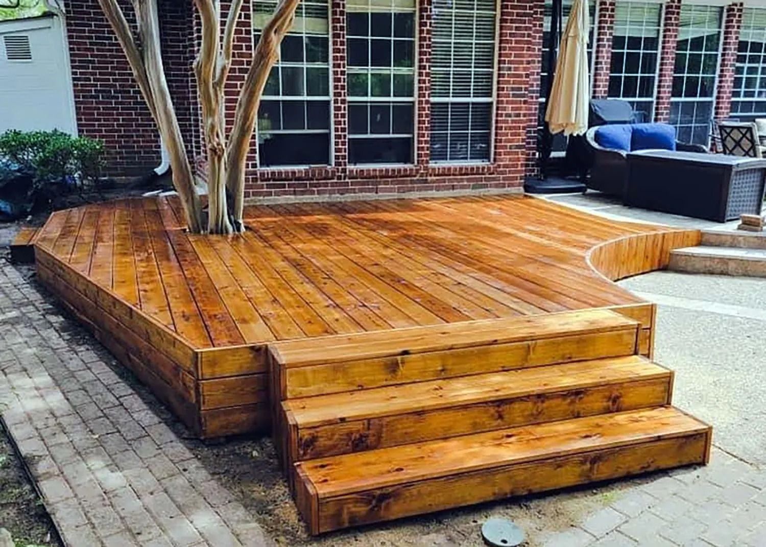 Wooden deck with steps in a brick patio backyard beside a tree and outdoor seating
