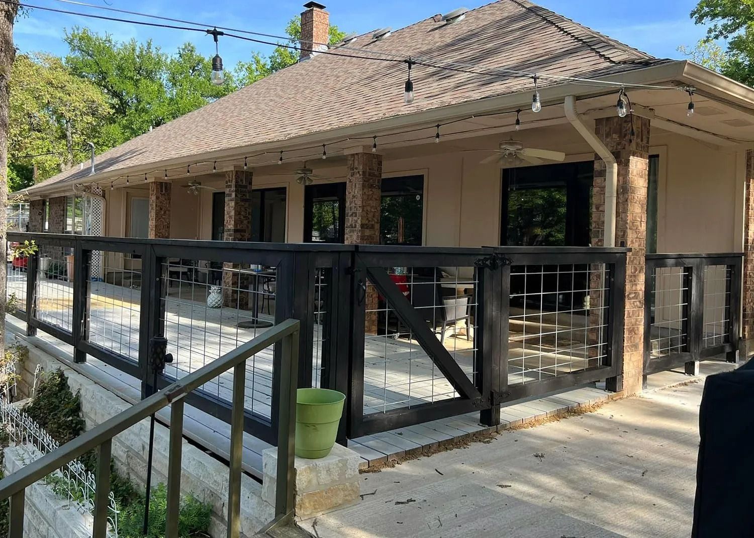 Covered patio with black railings on a beige house, shaded by a tiled roof and surrounded by greenery
