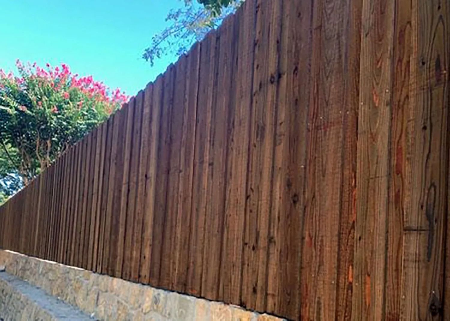 Long wooden privacy fence beside a stone wall, with pink flowers and blue sky above
