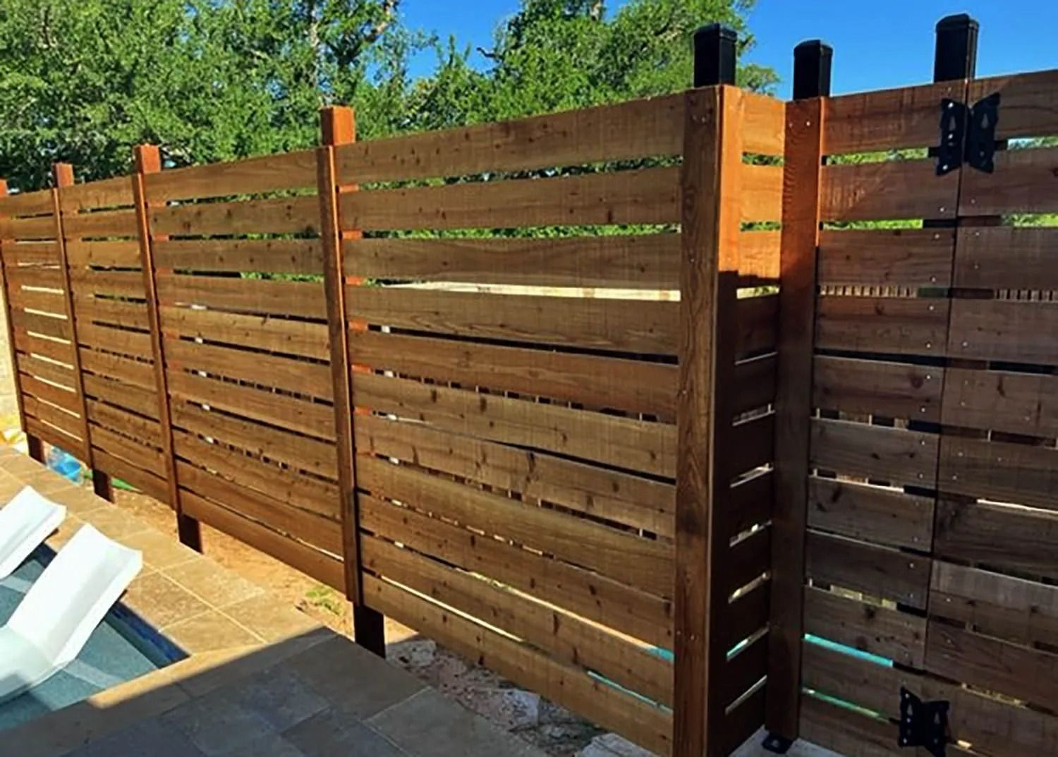 Brown wooden privacy fence along a patio, with trees and blue sky in the background

