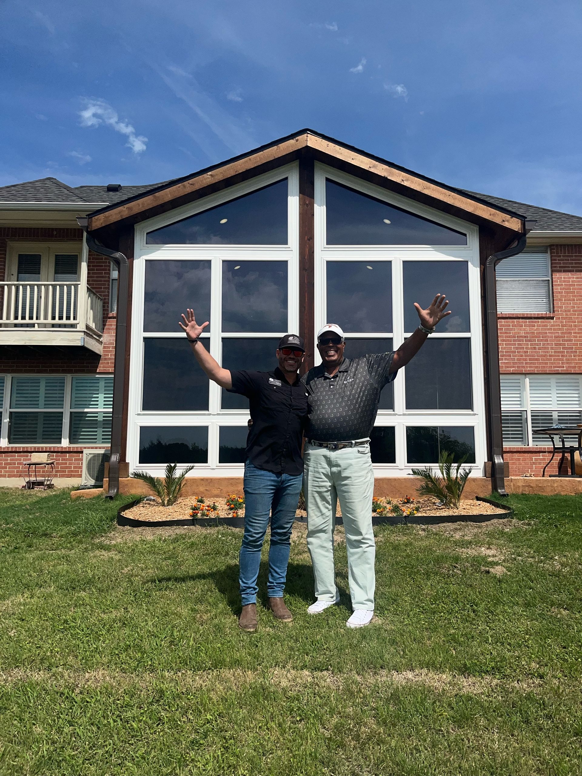 Two people posing in front of a large house with raised arms and a blue sky behind them