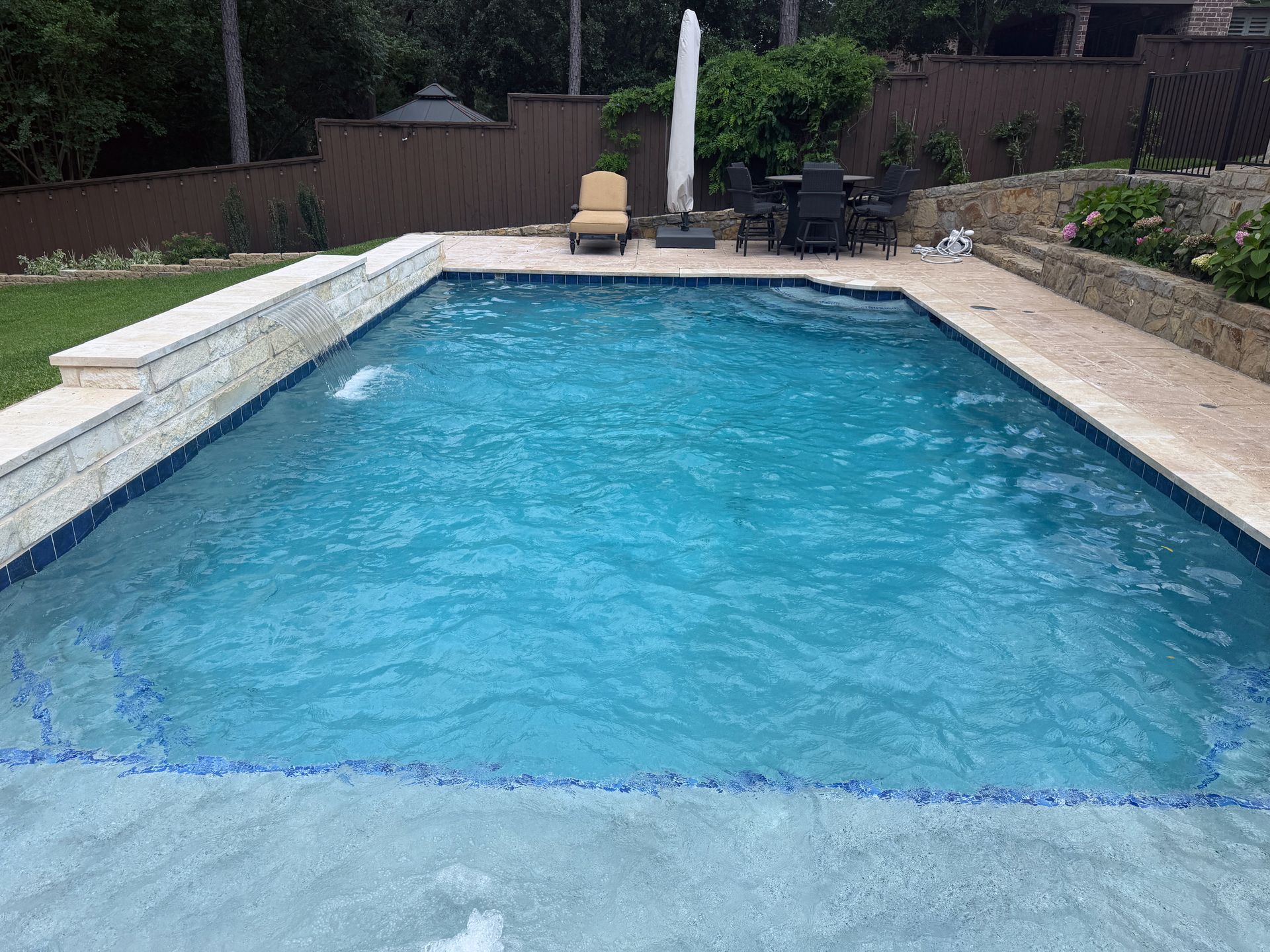 Backyard swimming pool with blue water, stone coping, and patio chairs in the background