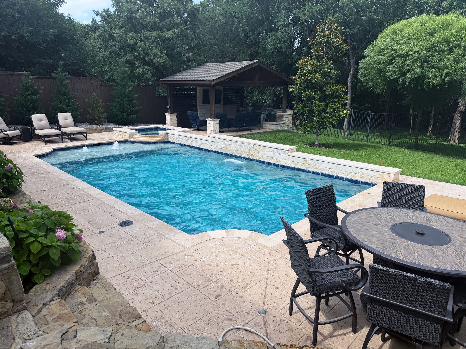 Backyard pool with stone patio, lounge chairs, round table, and a shaded pool house near trees