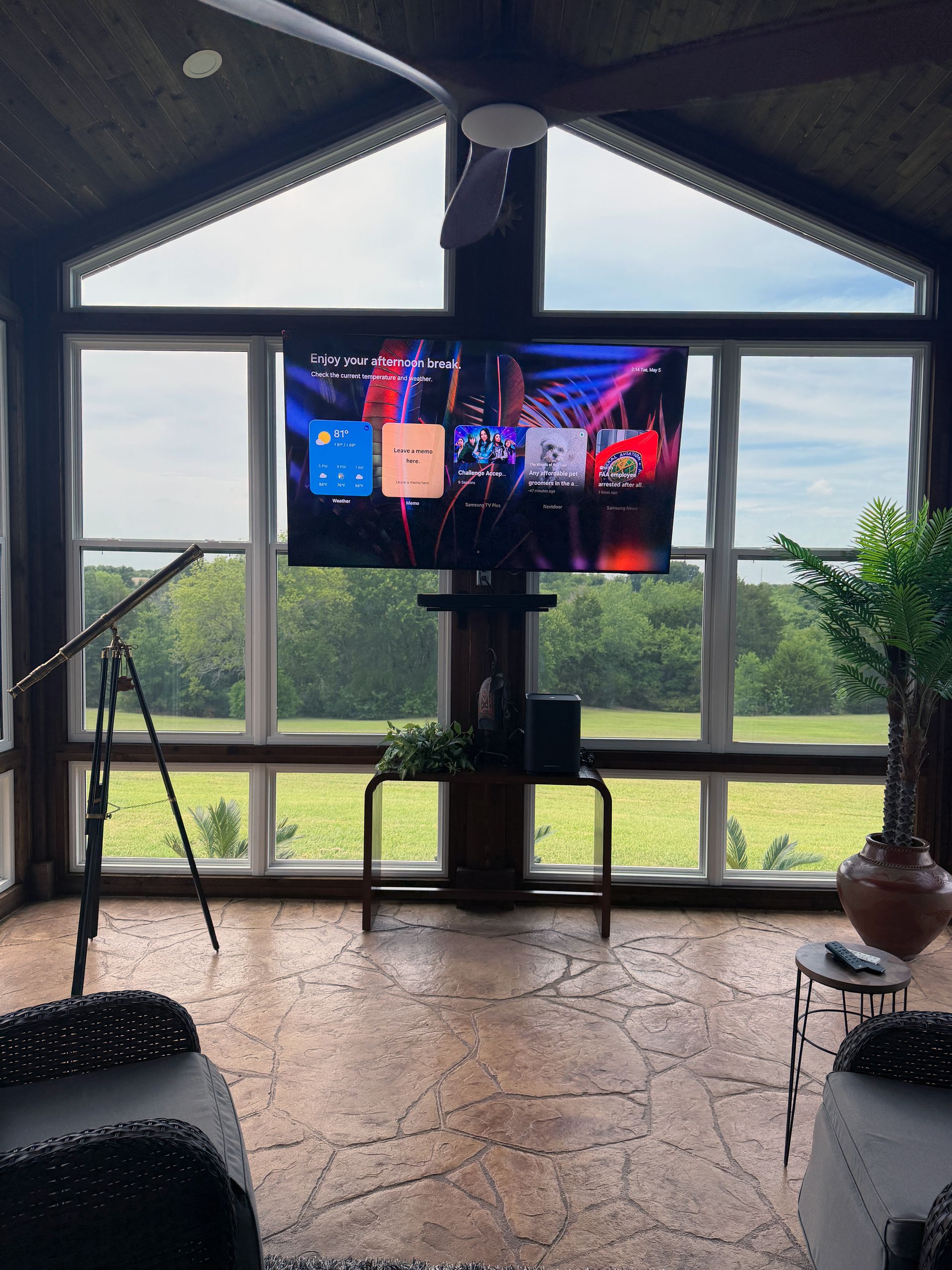 Living room with a TV on a stand, large windows, and a view of greenery outside.