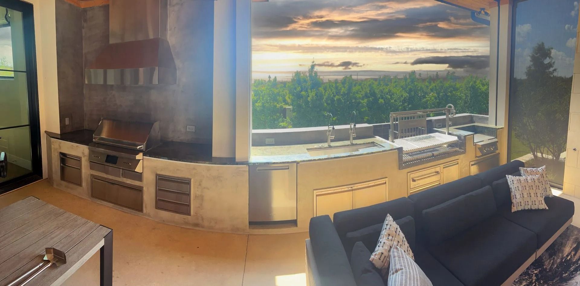 Modern kitchen and living room with large window, sunset view, and dark sofa near the counter.