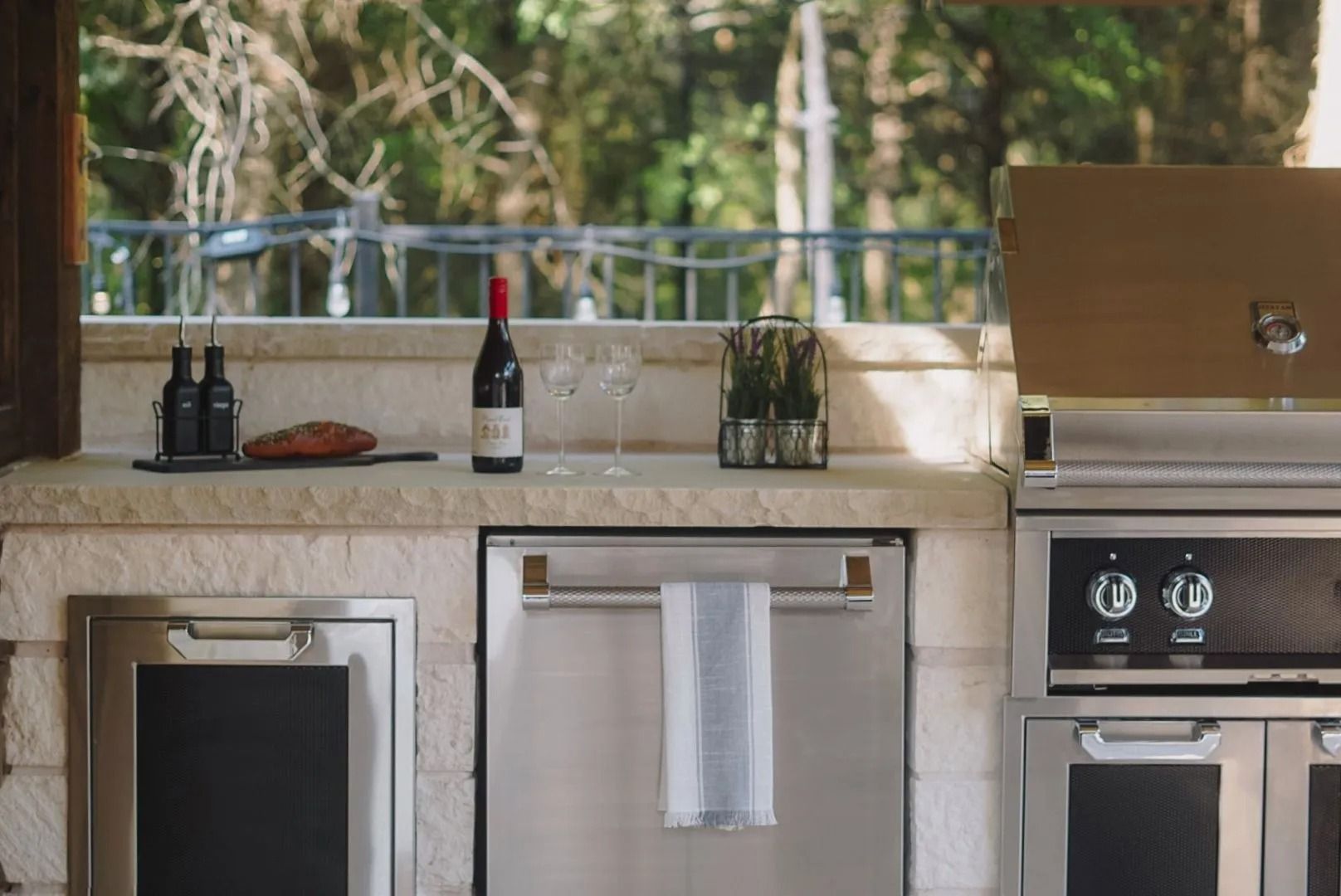 Outdoor kitchen countertop with wine bottle, glassware, towel, and built-in appliances by a window