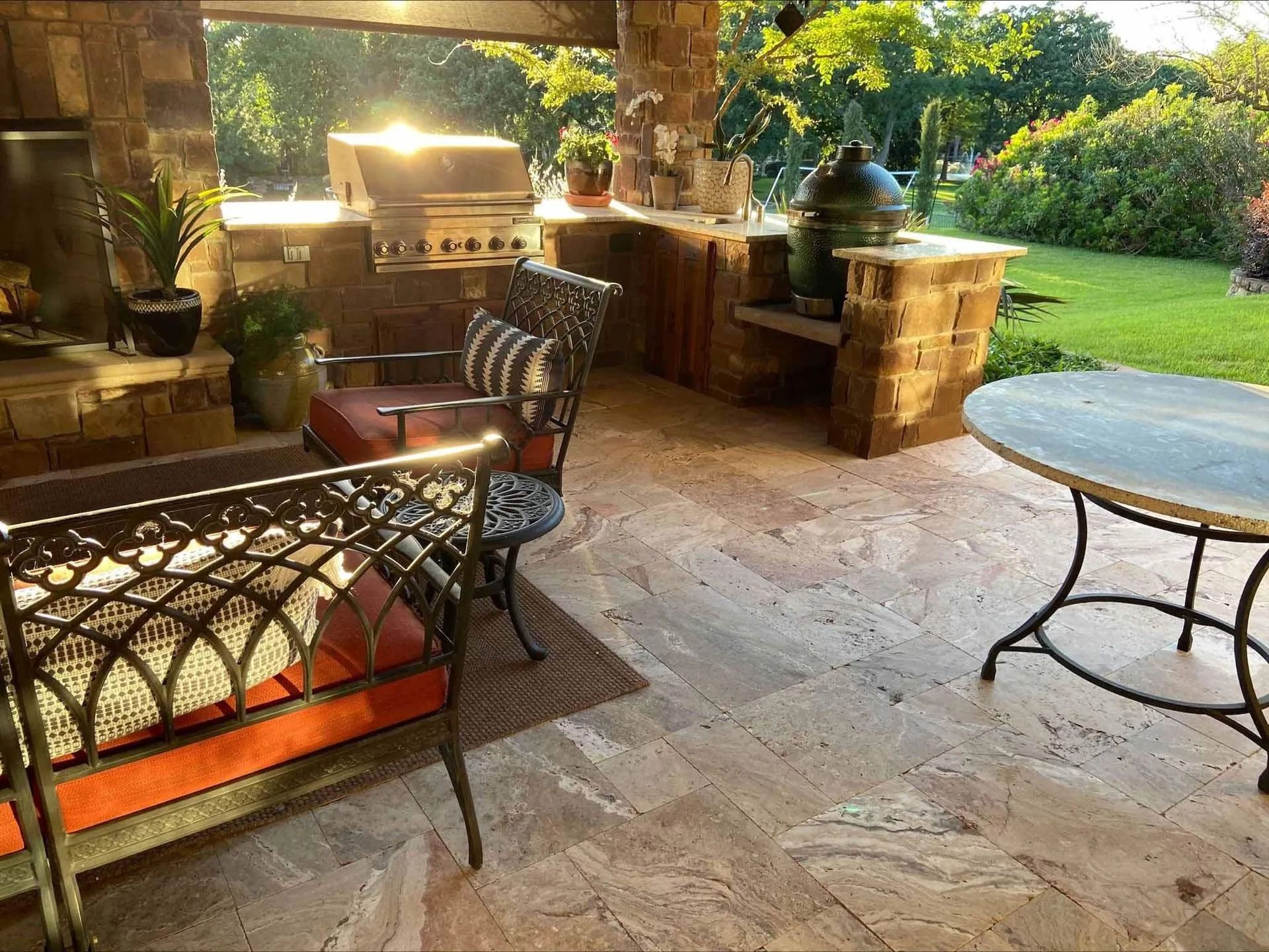 Outdoor patio with stone floor, grill, metal chairs, and round table beside a sunlit garden