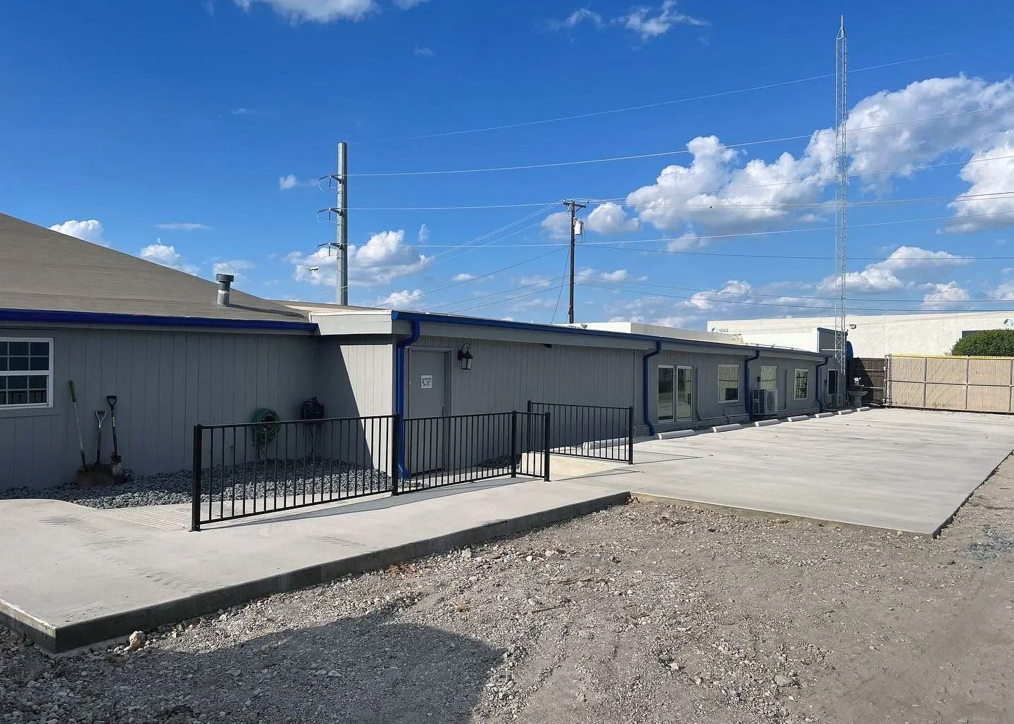 Single-story gray building with a fenced walkway and gravel lot under a blue sky
