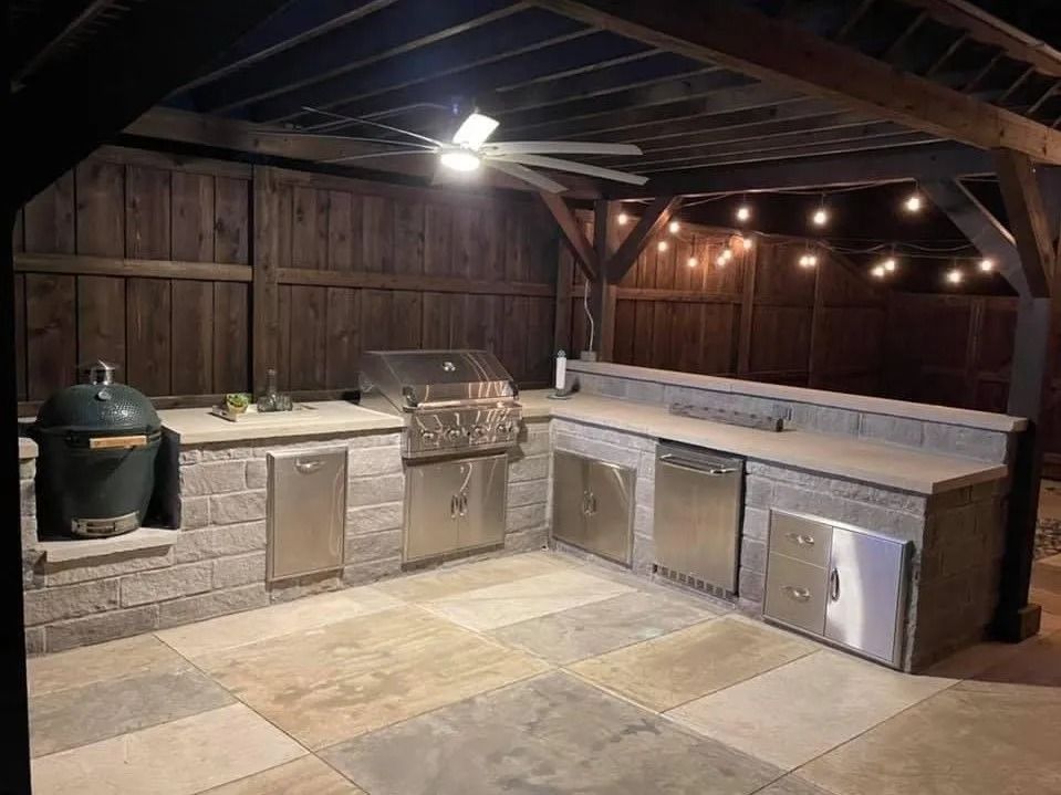 Outdoor patio kitchen with grill, sink, and stainless steel cabinets under a wooden pergola at night