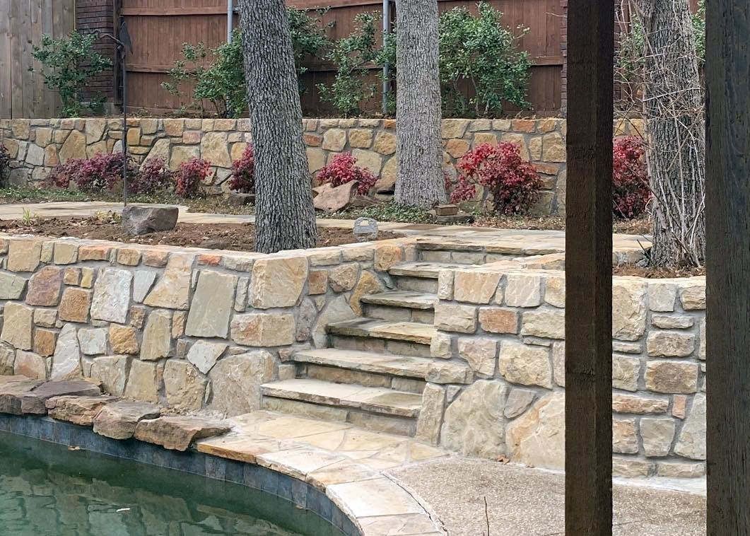 Stone steps and retaining wall beside a pool in a landscaped yard with trees and shrubs