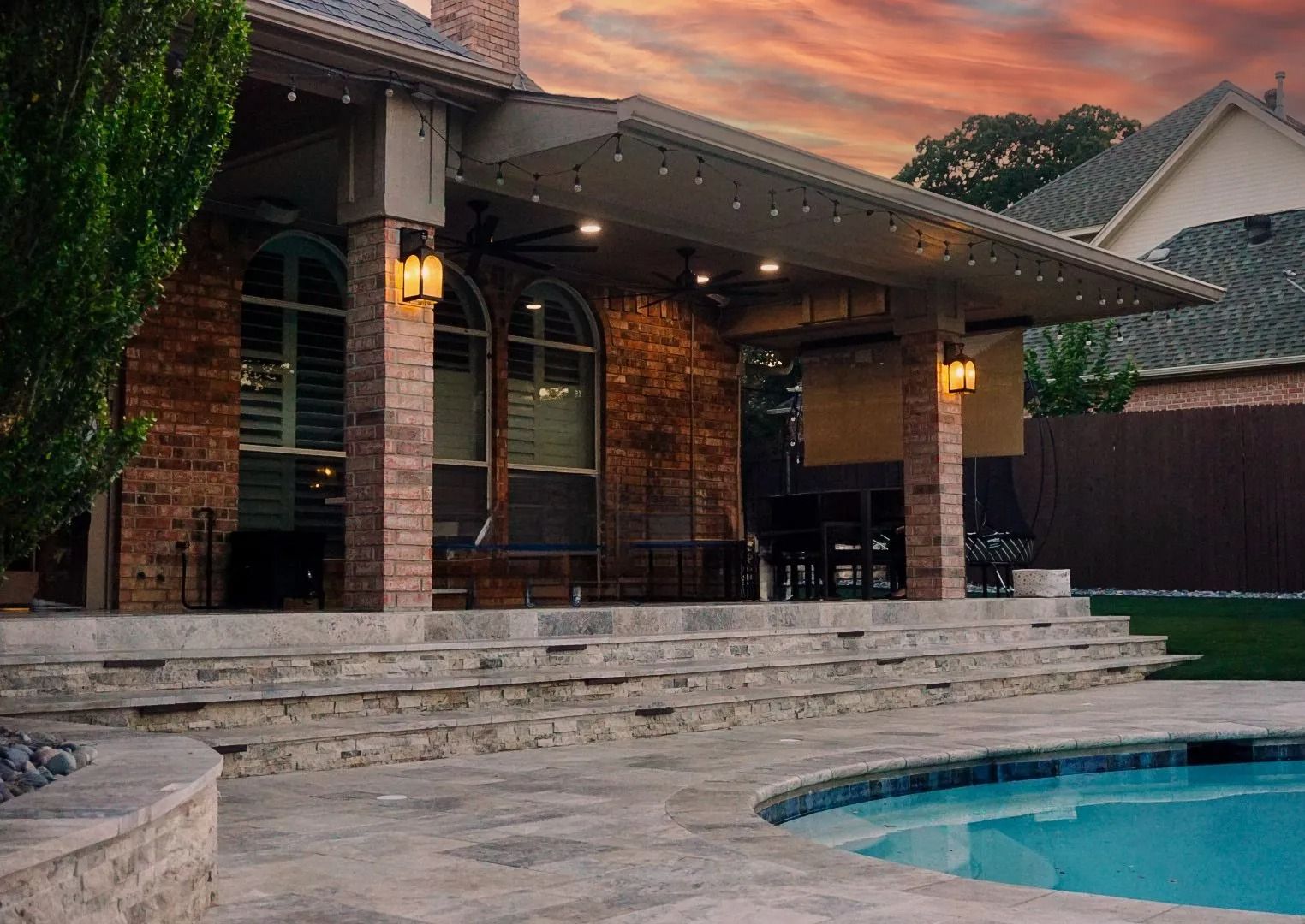 Backyard patio at dusk with stone steps, brick columns, warm lights, and a swimming pool beside the house