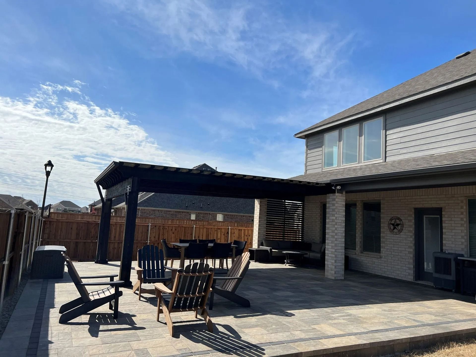 Back patio with a black pergola, patio chairs, and a two-story house under a blue sky
