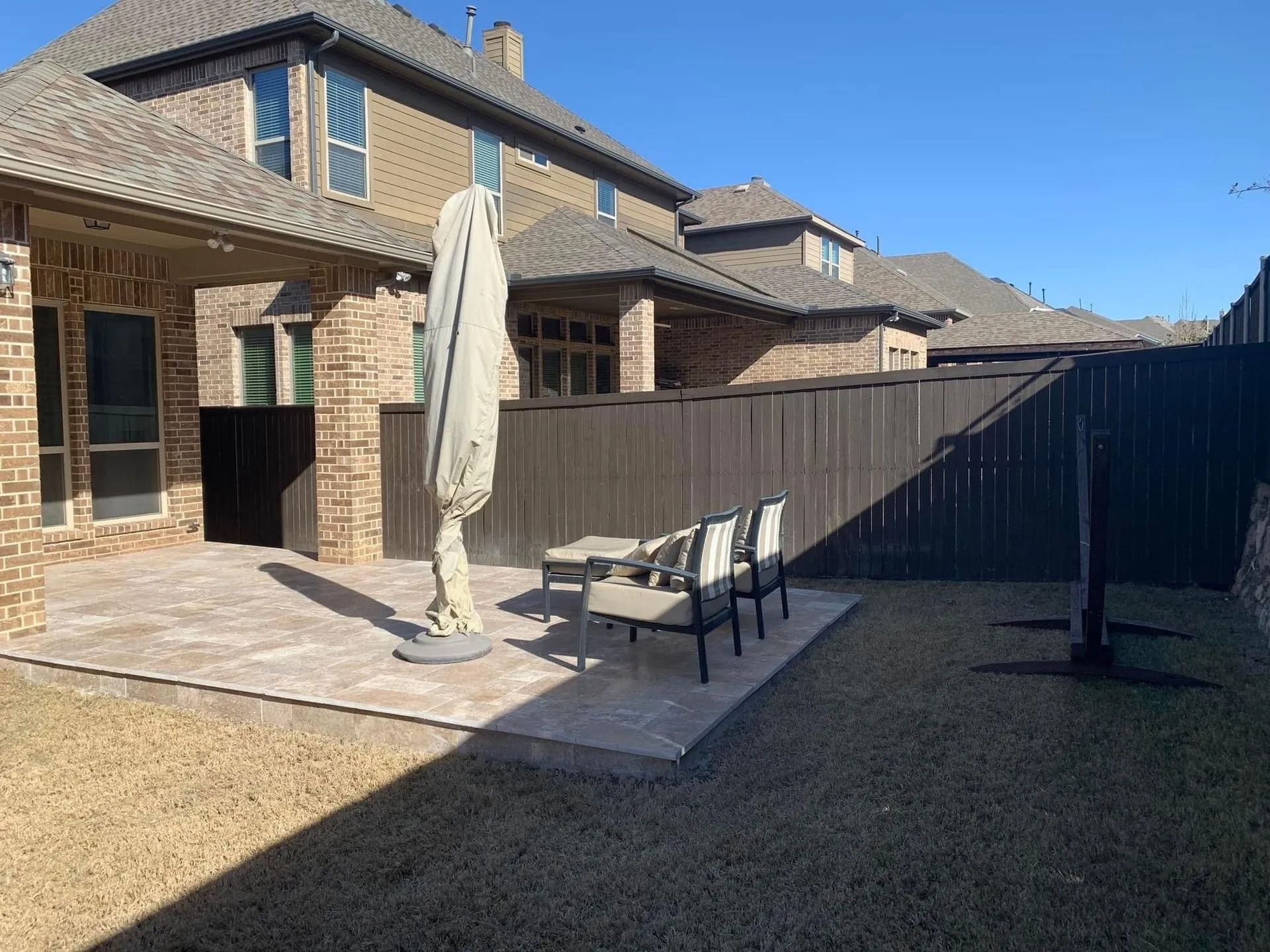 Backyard patio beside a brick house, with outdoor table, chairs, umbrella, and fence under clear blue sky