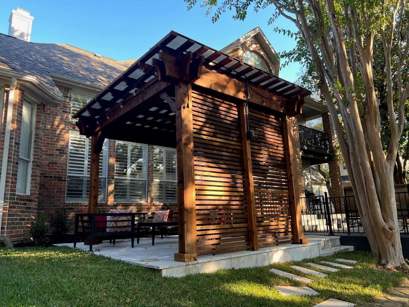 Wooden patio pavilion beside a brick house, with slatted walls and trees in the yard

