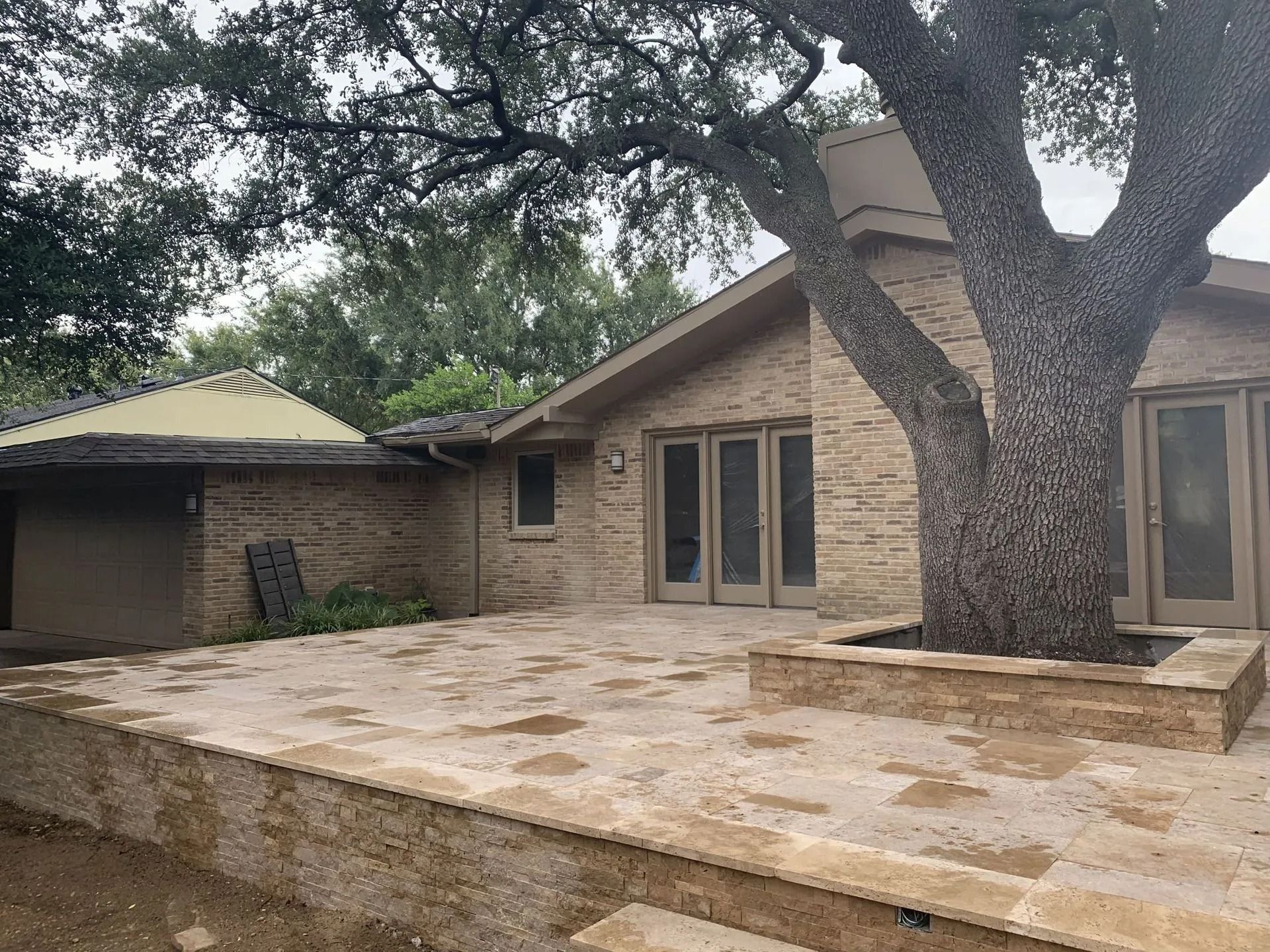 Stone patio and large tree in a backyard beside a beige brick house with a covered porch