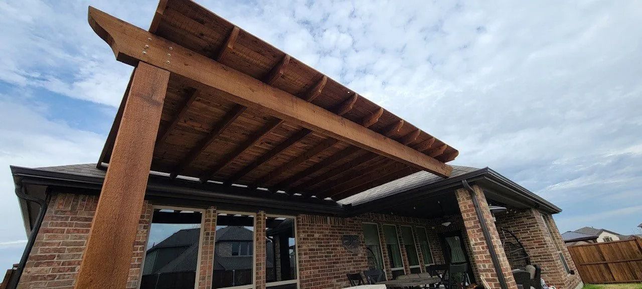 Wooden pergola roof structure under construction outside a stone building, viewed from below.